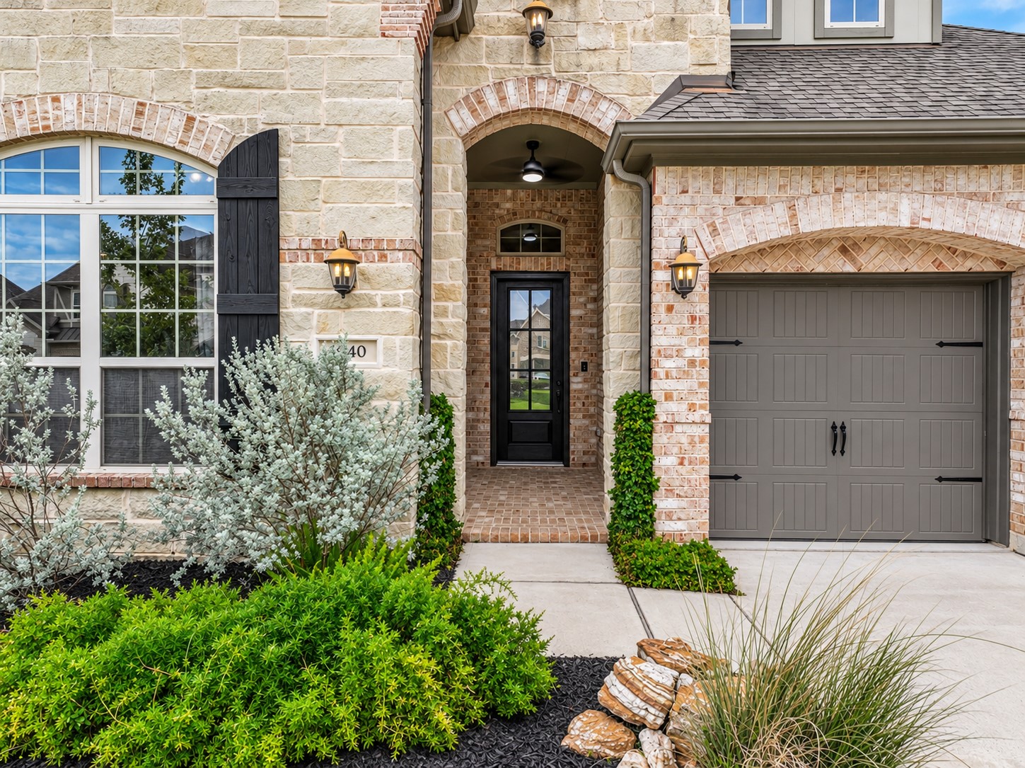 A closer look at the home's welcoming covered entry, framed by Austin limestone, decorative brick arches & attractive landscaping. Notice the carriage-style garage door, dark-stained wood shutter & brick-paved porch tucked beneath the impressive arched entrance.
