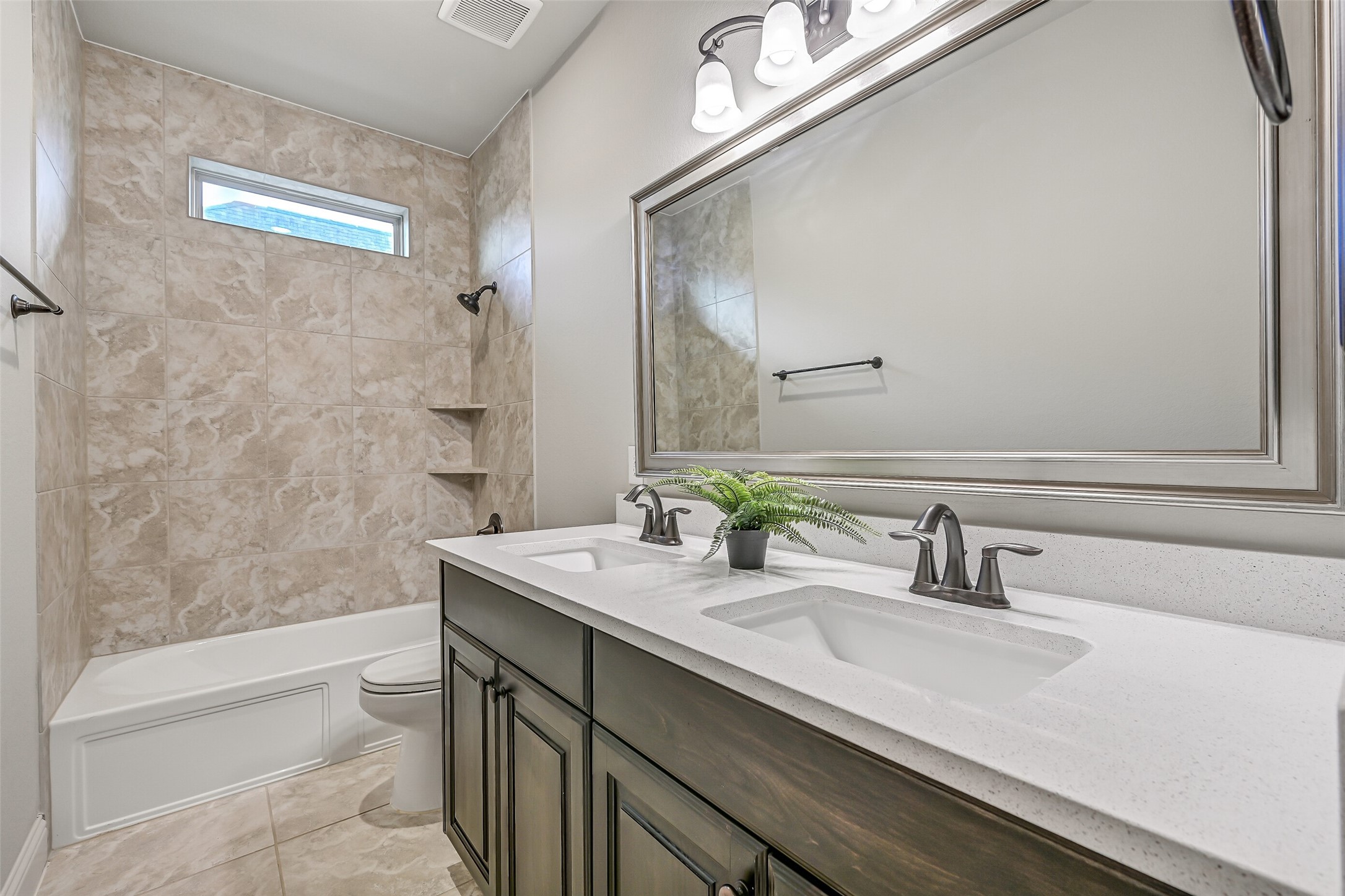 Situated between two secondary bedrooms, this full bath features a quartz countertop with dual rectangular sinks, stained wood cabinetry, a large framed mirror & a tub/shower combination with a tiled surround. A high-set window brings in natural light.