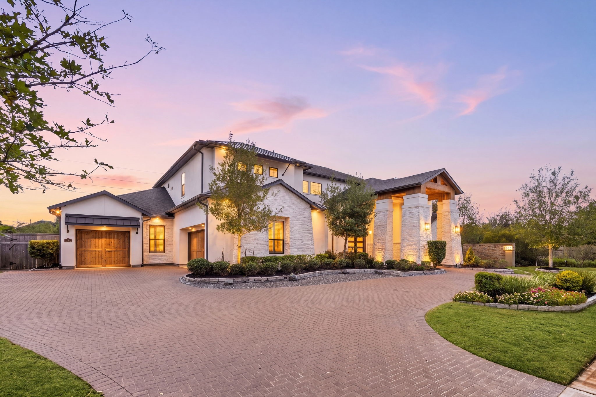 Impressive front elevation featuring an expansive driveway, mature landscaping, and a welcoming entrance. The pave stone circle drive adorn the entrance along with beautiful elevation including metal roof accents, stone and stucco architecture, and stained garage doors.