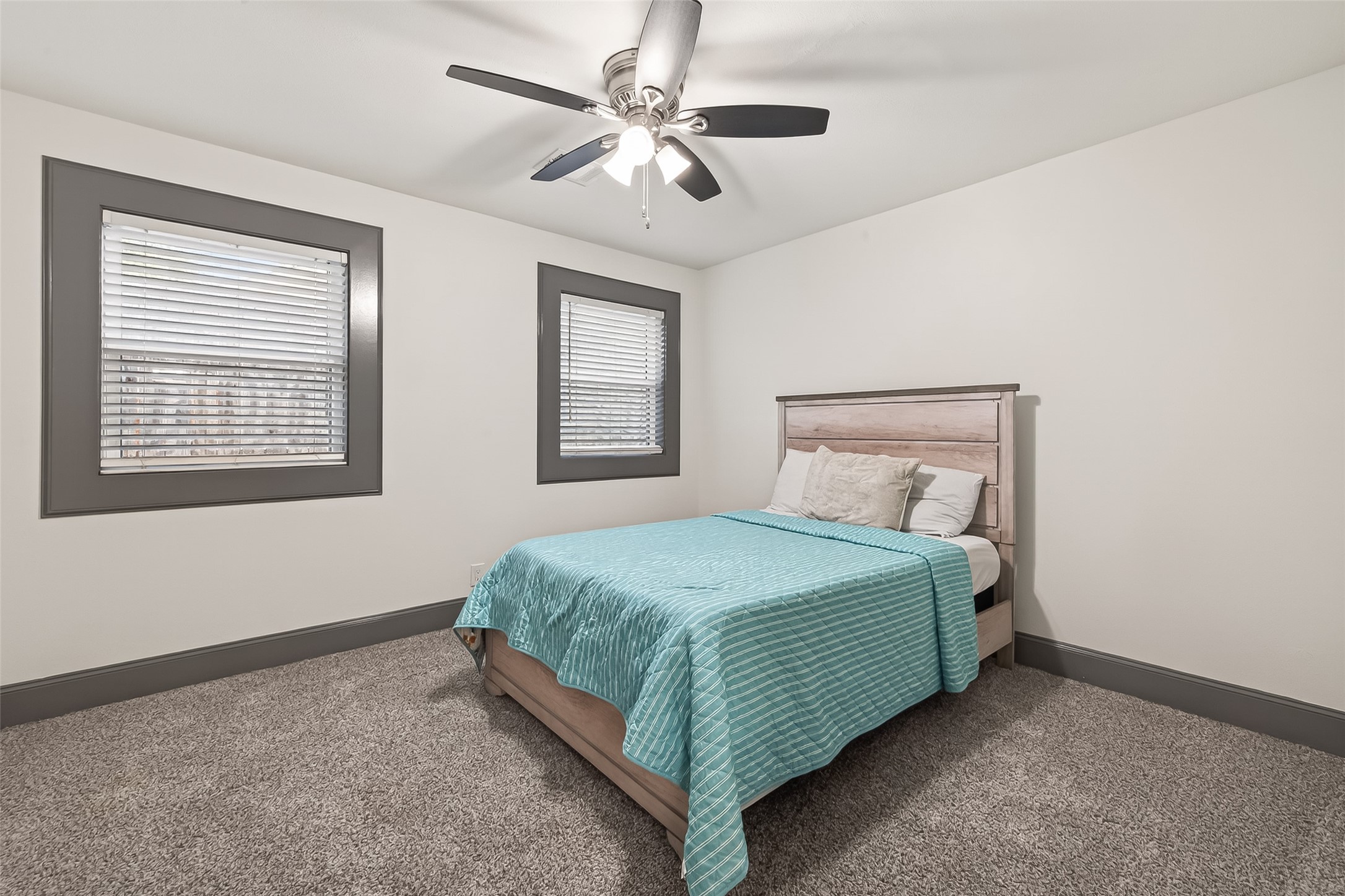 This bedroom features neutral decor with gray trim, two windows for natural light, plush carpet, and a ceiling fan for comfort.