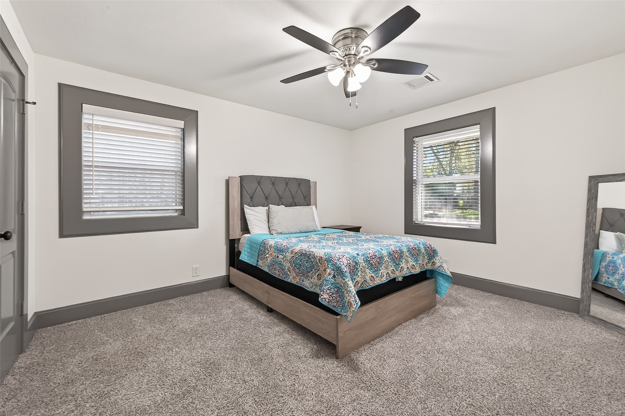 This bedroom is cozy, with a carpeted floor with a modern ceiling fan and two windows for natural light.
