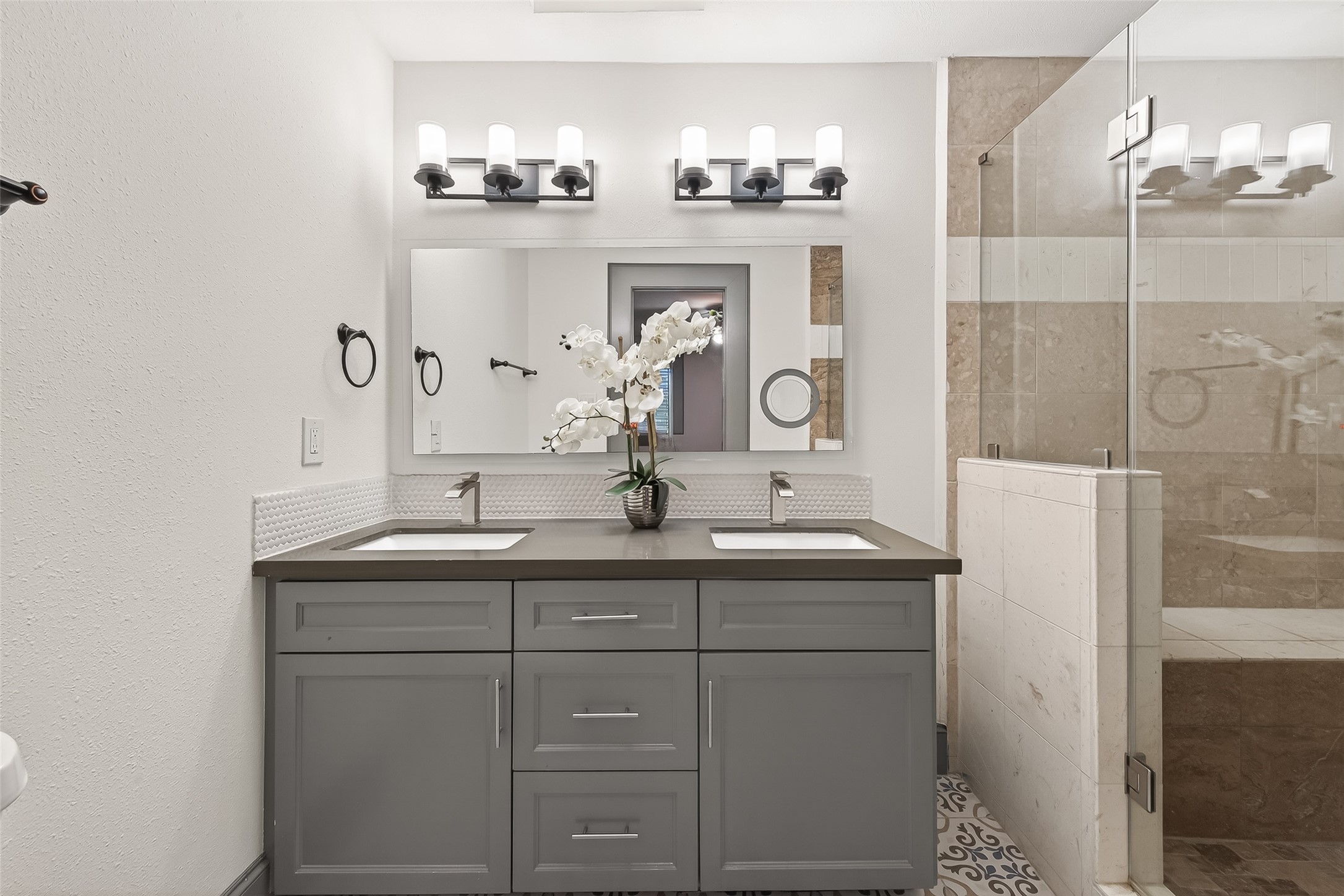 This bathroom is a modern design with a double vanity, sleek gray cabinets, and elegant lighting fixtures.