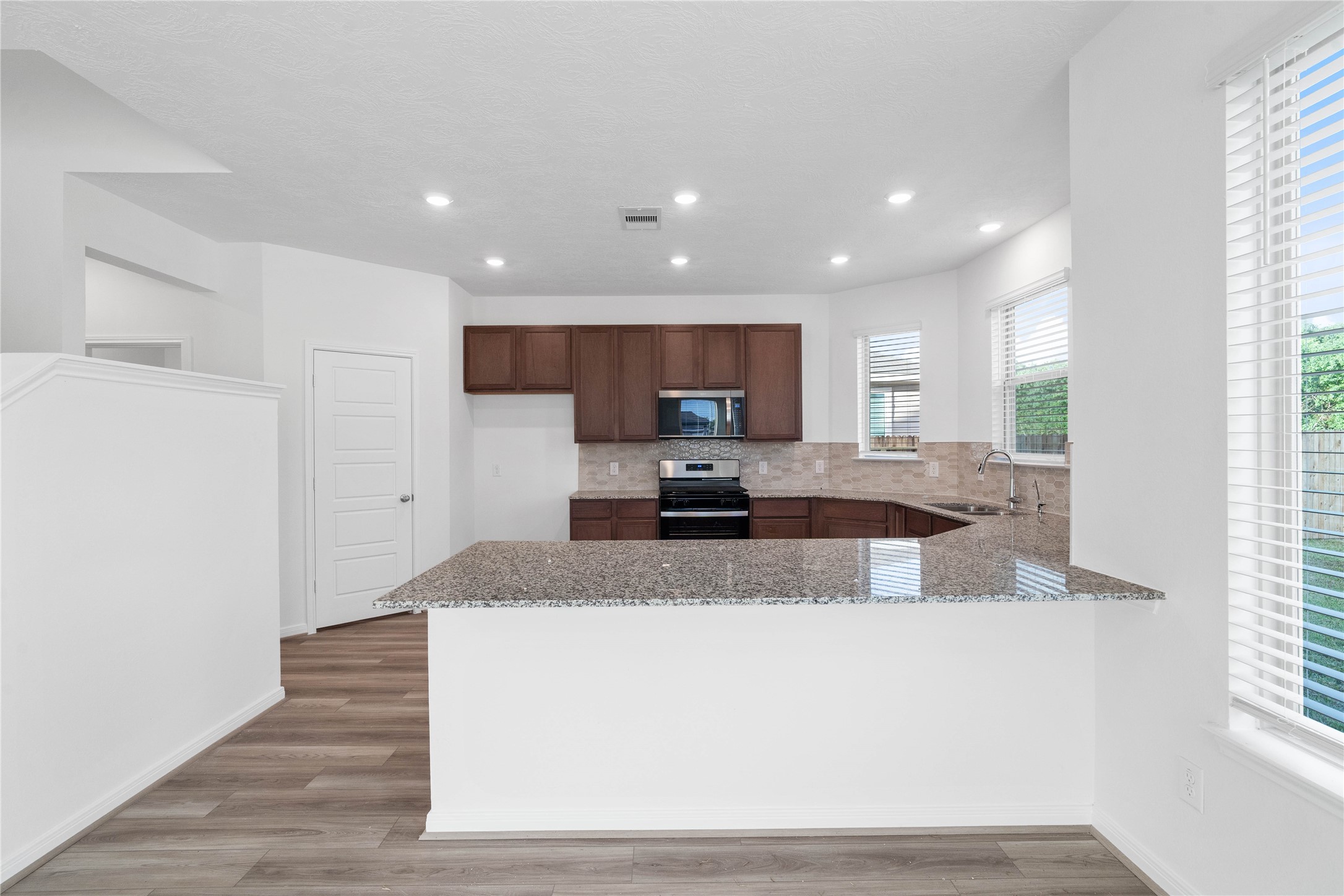 The kitchen features rich ginger stained wood cabinetry, granite countertops, and a pearl colored picket mosaic backsplash that adds subtle texture and style. Warm wood look flooring and natural light create an inviting space for everyday cooking and gathering.