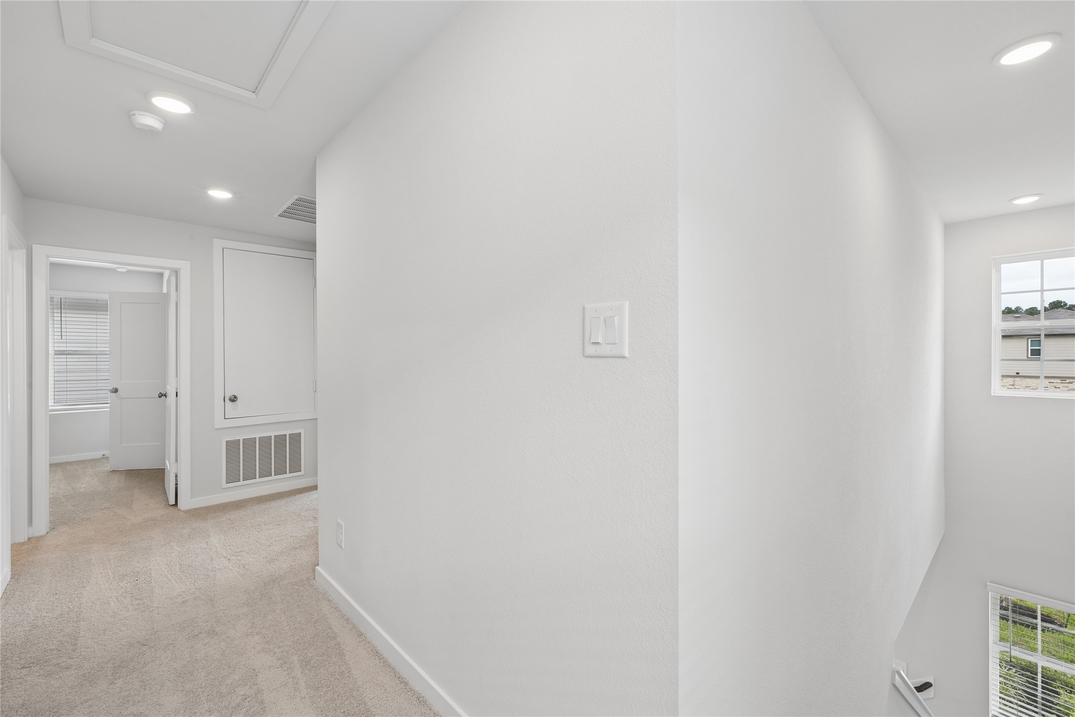 Bright and inviting hallway with plush carpeting, recessed lighting, and ample natural light from large windows, leading to a cozy room.