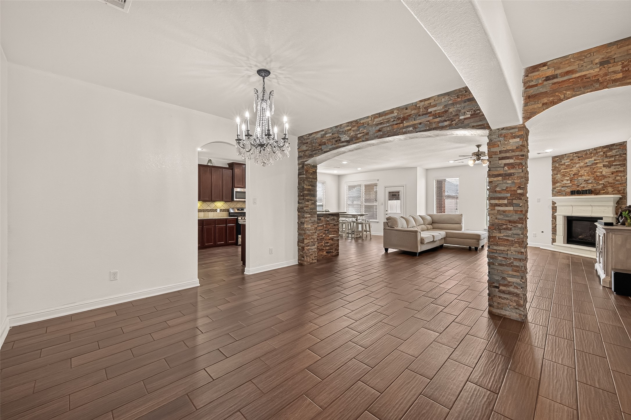Elegant formal dining room flows into the living area, featuring wood-look tile flooring, a stone fireplace, and a stylish chandelier, with seamless access to a modern kitchen with rich cabinetry.