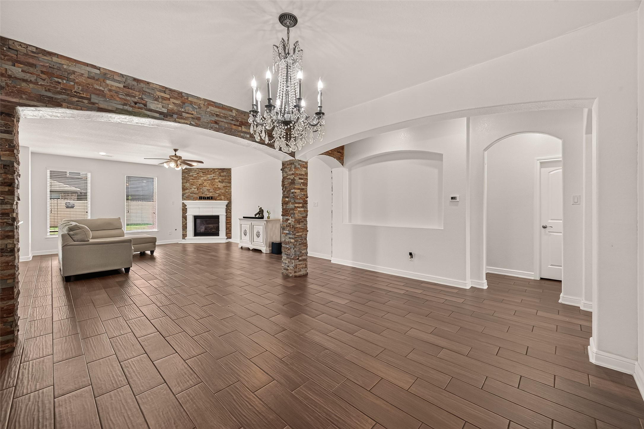 Elegant living space featuring rich wood-like flooring, a cozy fireplace, and a stunning chandelier. The room is accentuated by stone columns and archways, offering a blend of modern and classic charm.
