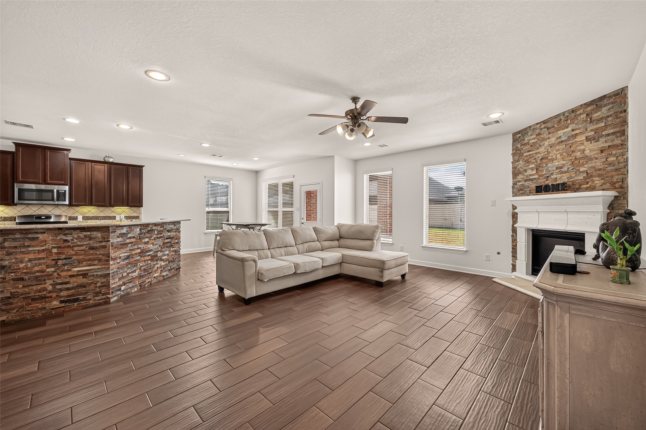 Spacious open concept living area with a cozy stone fireplace, elegant wood-look tile flooring, and a modern kitchen featuring rich cabinetry and a stone-accented breakfast bar.