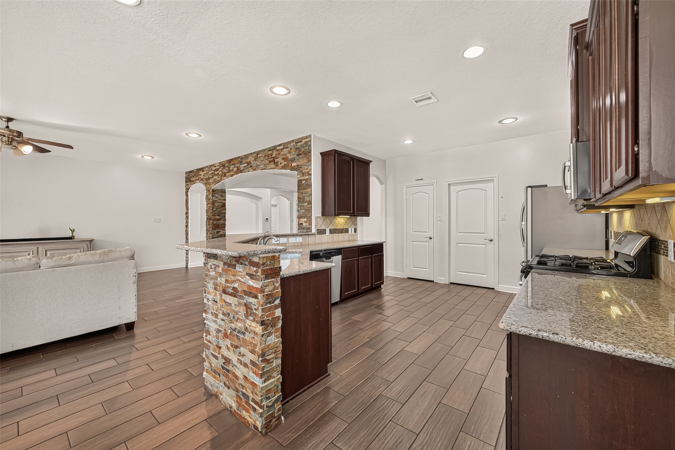 Spacious modern kitchen featuring dark wood cabinetry, sleek granite countertops, and a striking stone accent wall, flowing seamlessly into a cozy living room.