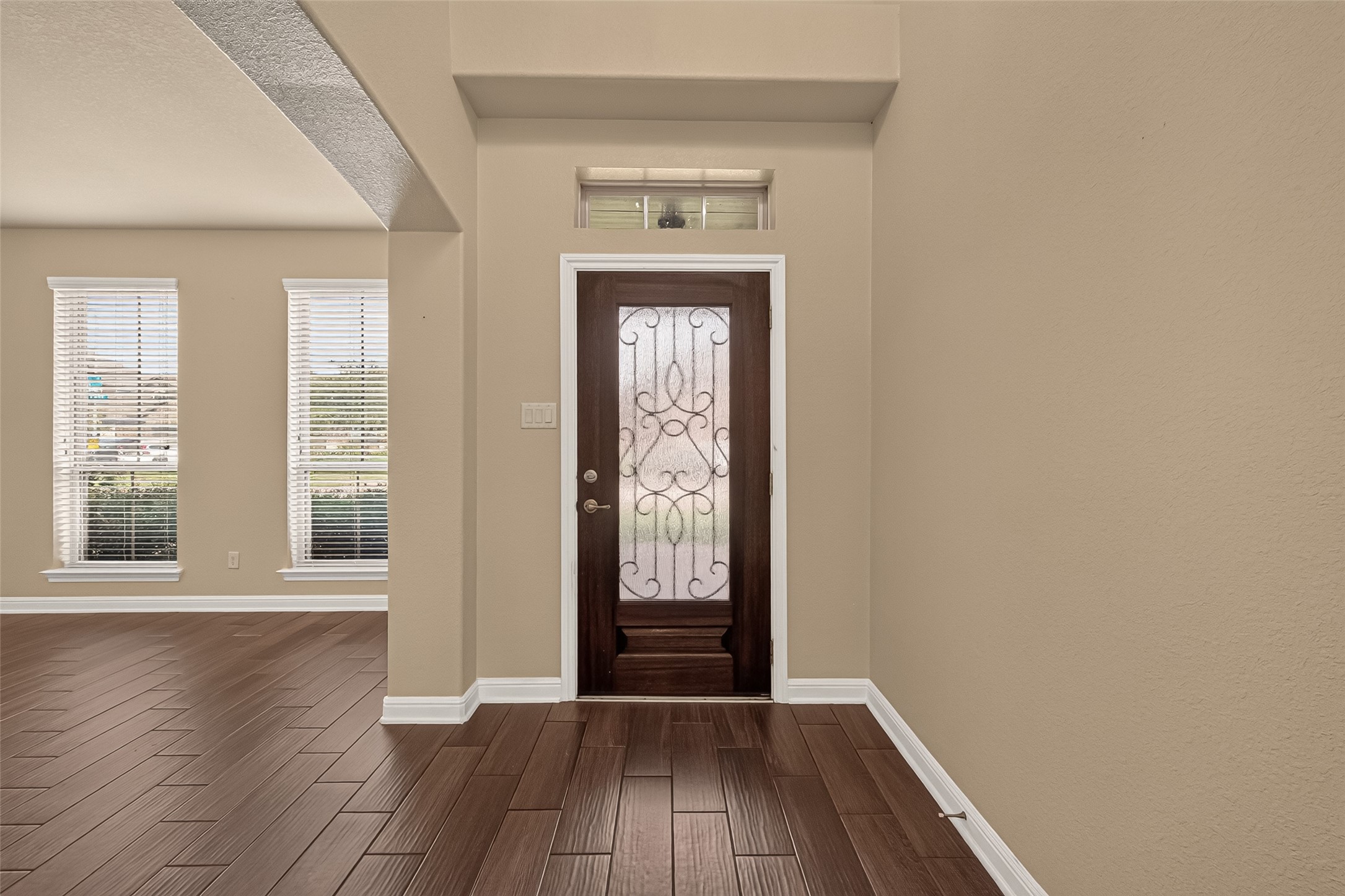 Inviting entryway with elegant wood flooring and a decorative glass front door, leading to a bright living area with ample natural light from large windows.
Elegant entryway featuring rich wood-look tile flooring and graceful archways, leading to a charming staircase with decorative iron balusters.