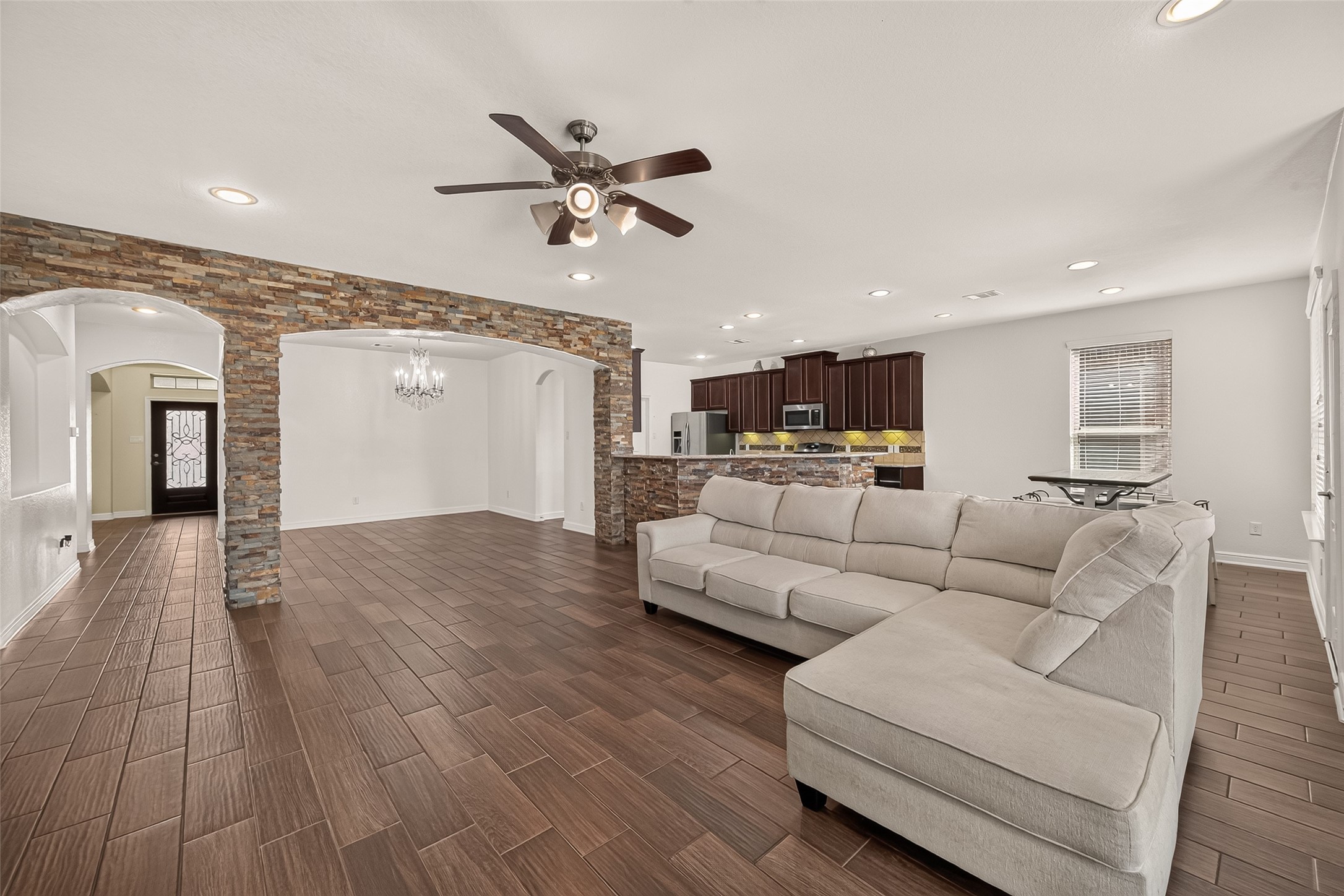 Spacious open-concept living area features elegant tile flooring, a modern ceiling fan, and a cozy sectional. The room flows seamlessly into a stylish kitchen with dark wood cabinetry and stainless steel appliances, while stone archways add a touch of rustic charm.