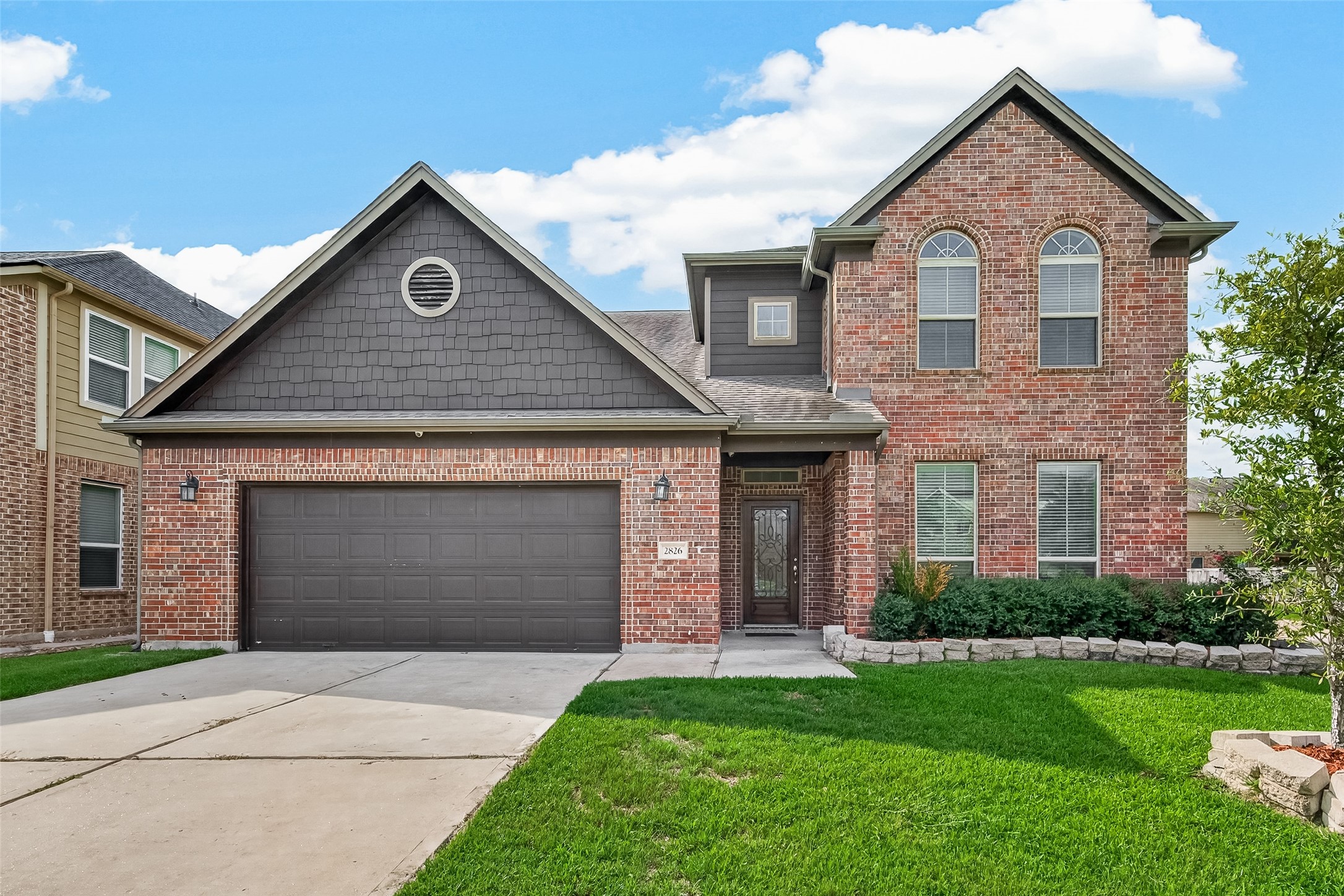 Charming two-story brick home featuring a spacious driveway, attached garage, and well-kept front lawn in a quiet neighborhood.