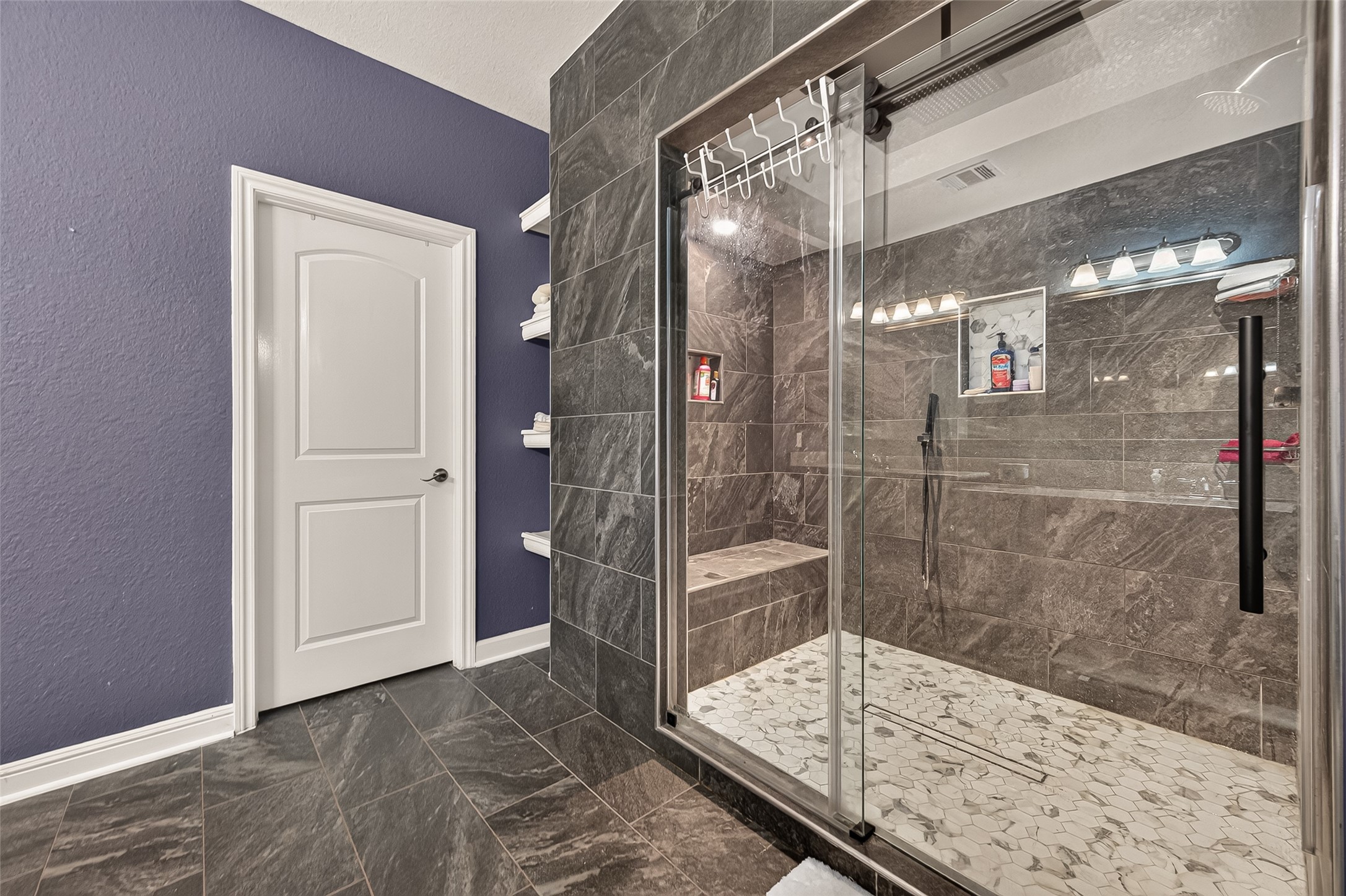 Luxurious bathroom featuring an expansive walk-in shower with frameless glass doors and sophisticated stone tilework, accented by a rich purple wall and convenient built-in shelving.