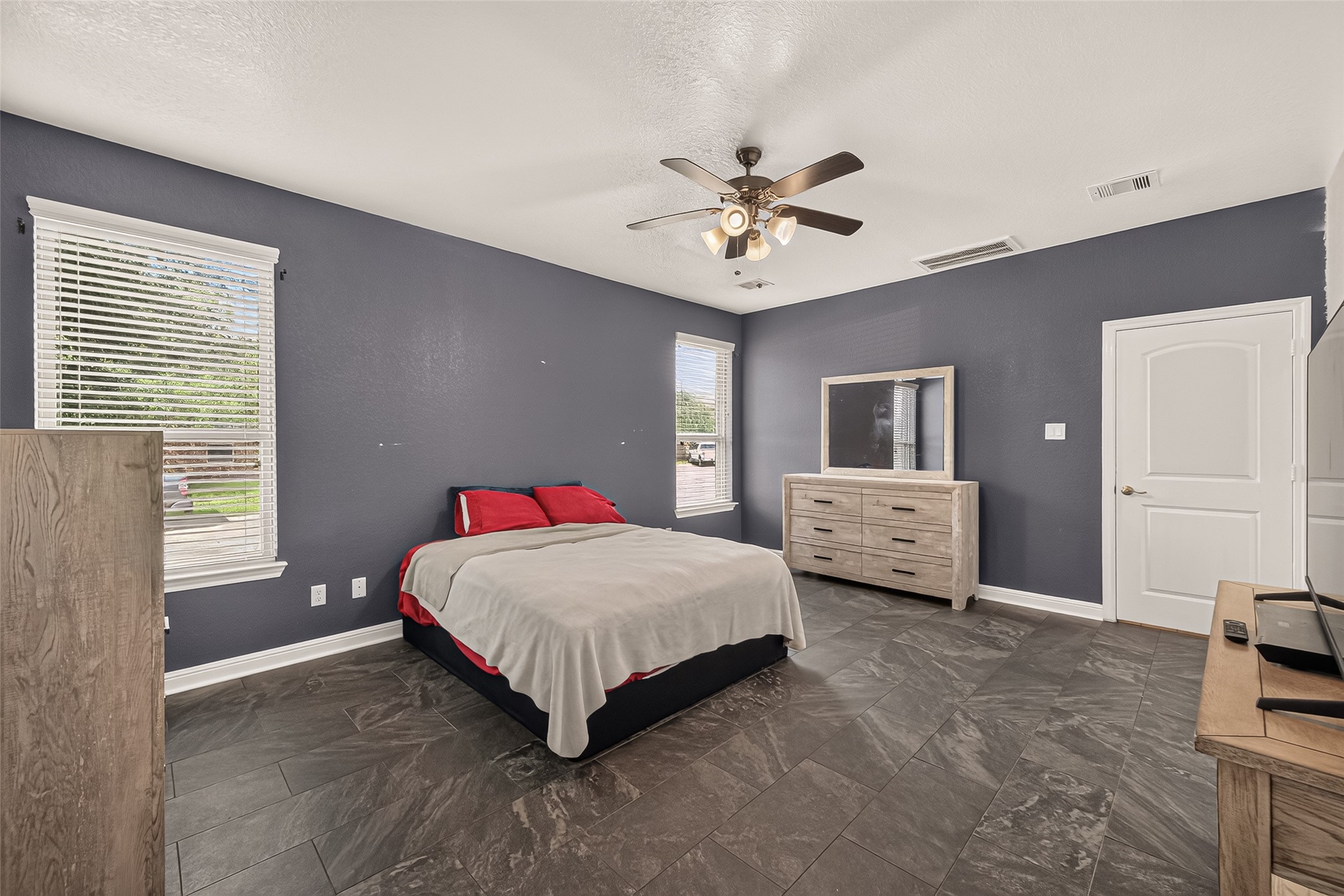 Spacious primary suite featuring sleek tile flooring, sophisticated dark walls, dual windows with abundant natural light, and a modern ceiling fan.