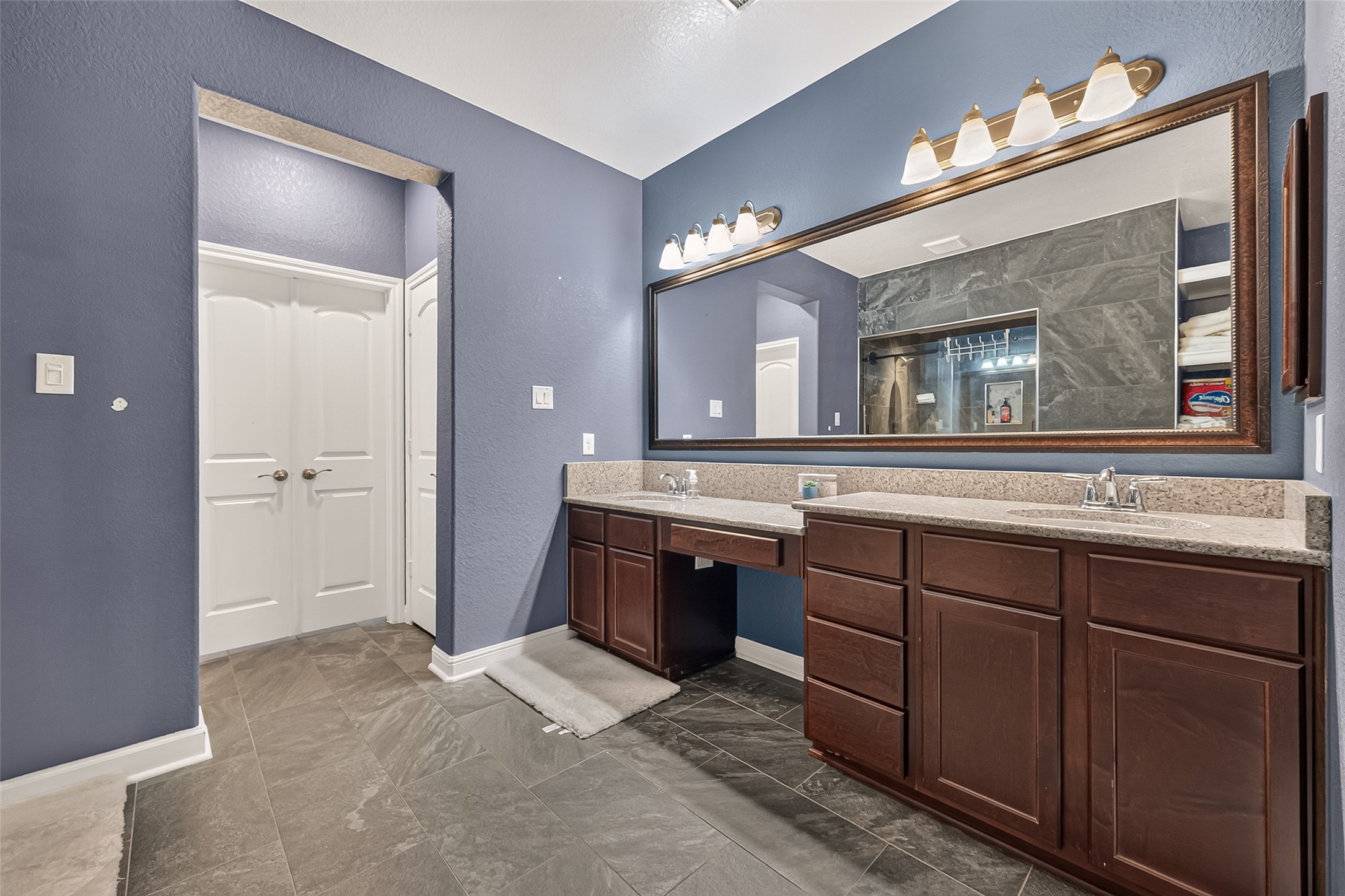 Refined primary bathroom showcasing dual vanities, finely crafted wood cabinetry, generous countertops, an oversized framed mirror, sophisticated lighting, and polished tile flooring.