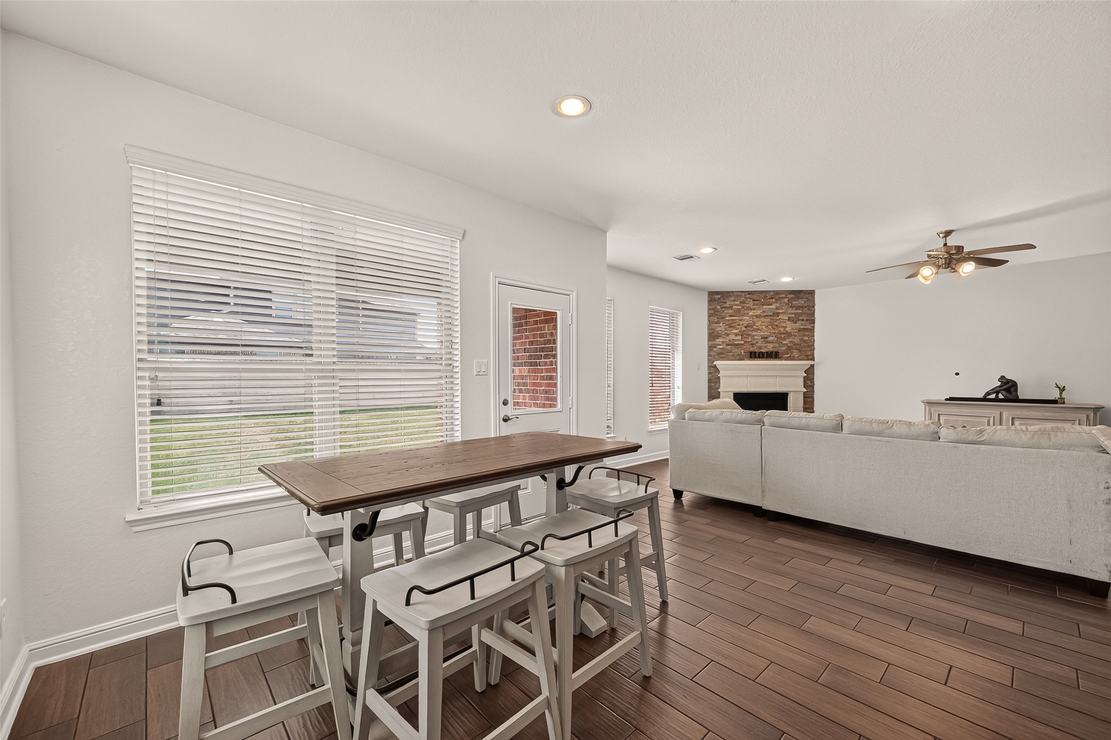 Spacious breakfast room opening to the living area, with elegant wood-look ceramic tile flooring, a cozy stone fireplace, and large windows that fill the space with natural light.