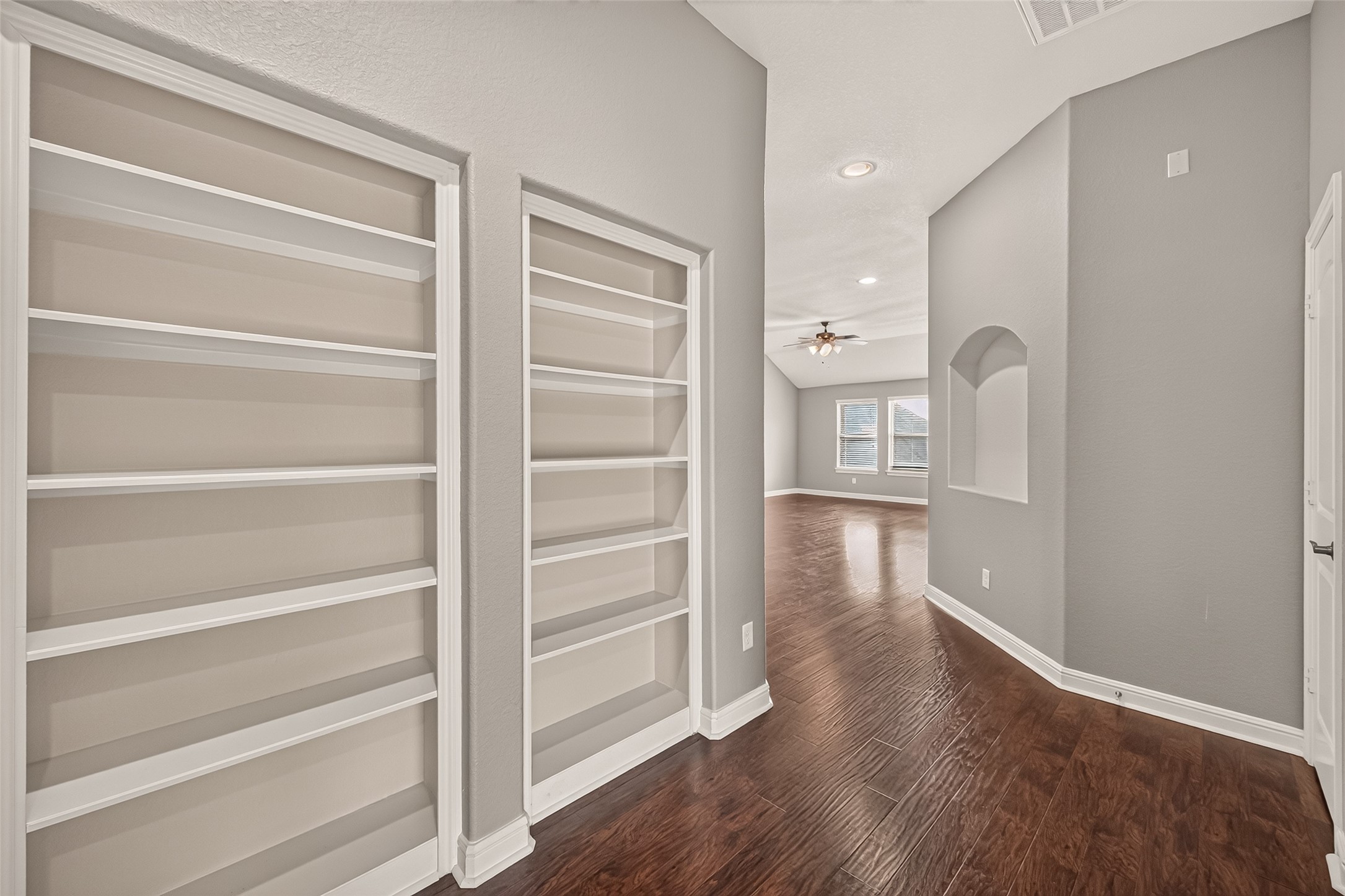 Elegant hallway with built-in shelving leads to a spacious living area, featuring rich hardwood floors, neutral walls, and abundant natural light.