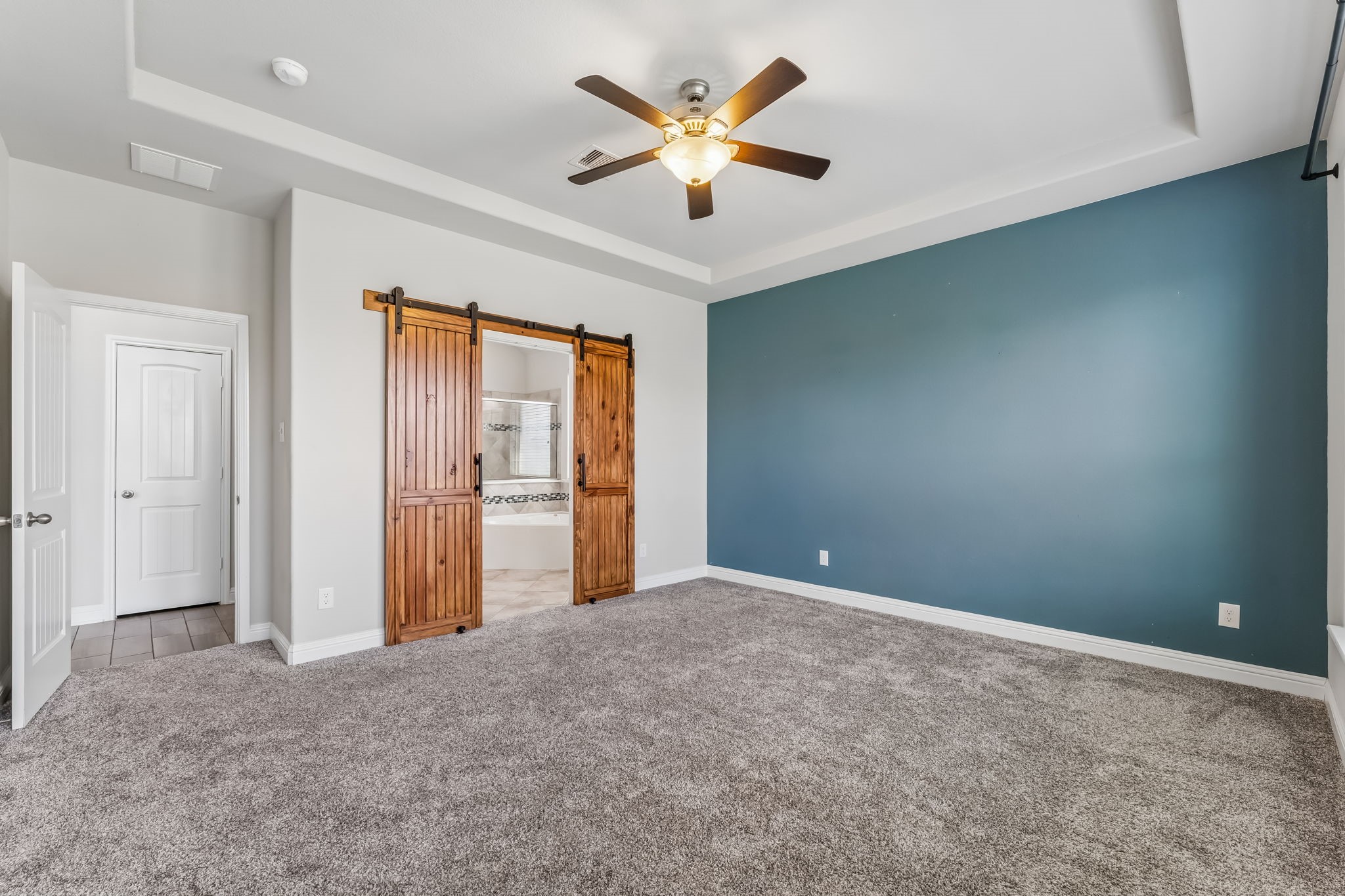 Spacious bedroom featuring a cozy carpet, a striking blue accent wall, and elegant wooden barn doors leading to an en-suite bathroom.