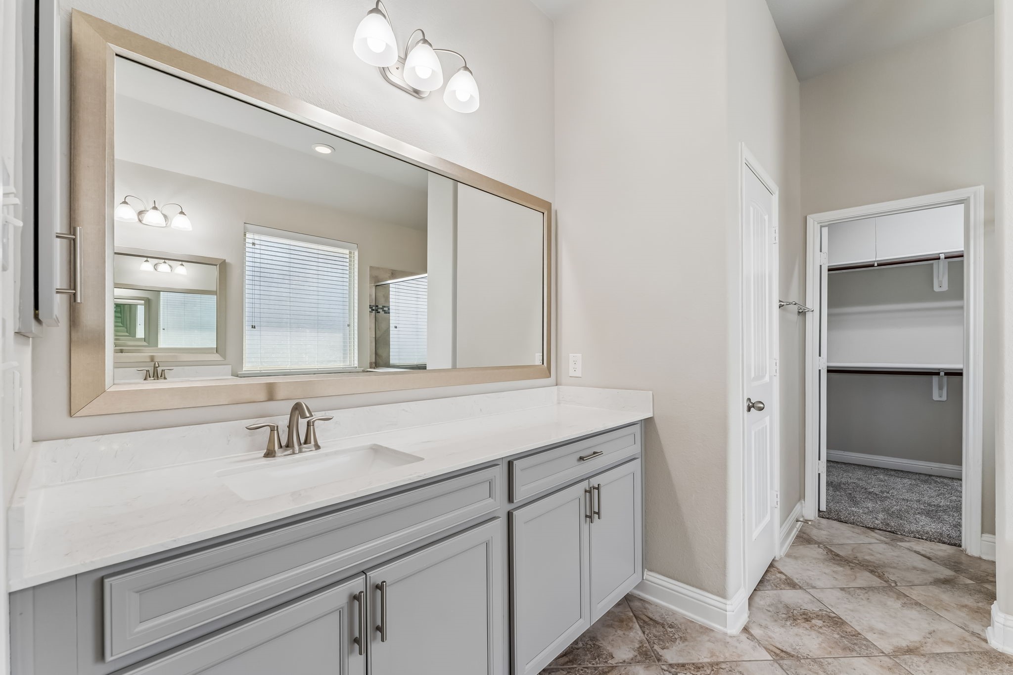 Elegant bathroom featuring a spacious vanity with a sleek countertop, large mirror, and modern lighting, leading into a roomy walk-in closet.