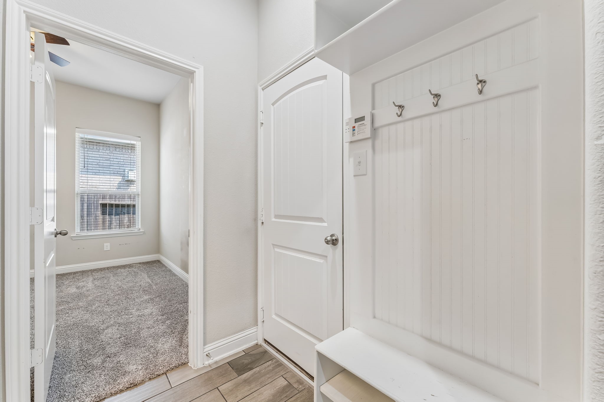 Charming entryway features a practical mudroom area with hooks and storage, leading into a cozy, carpeted room with a large window for natural light.