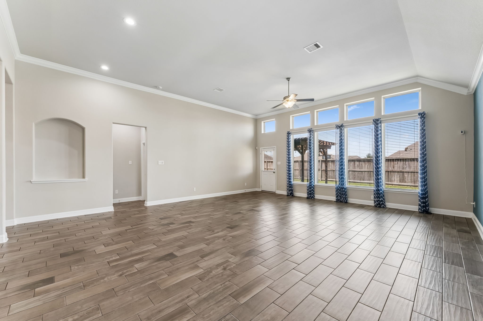 Spacious living area with high ceilings, abundant natural light from large windows, elegant tile flooring, and a ceiling fan for added comfort.