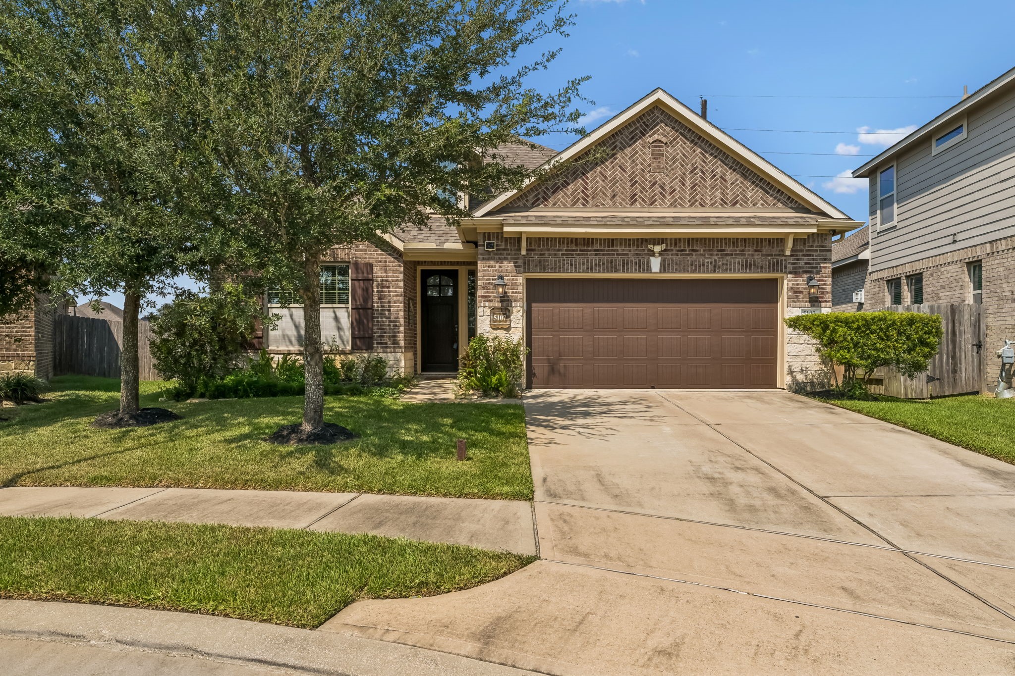 Charming brick home with a two-car garage, lush front lawn, and mature trees offering shade.