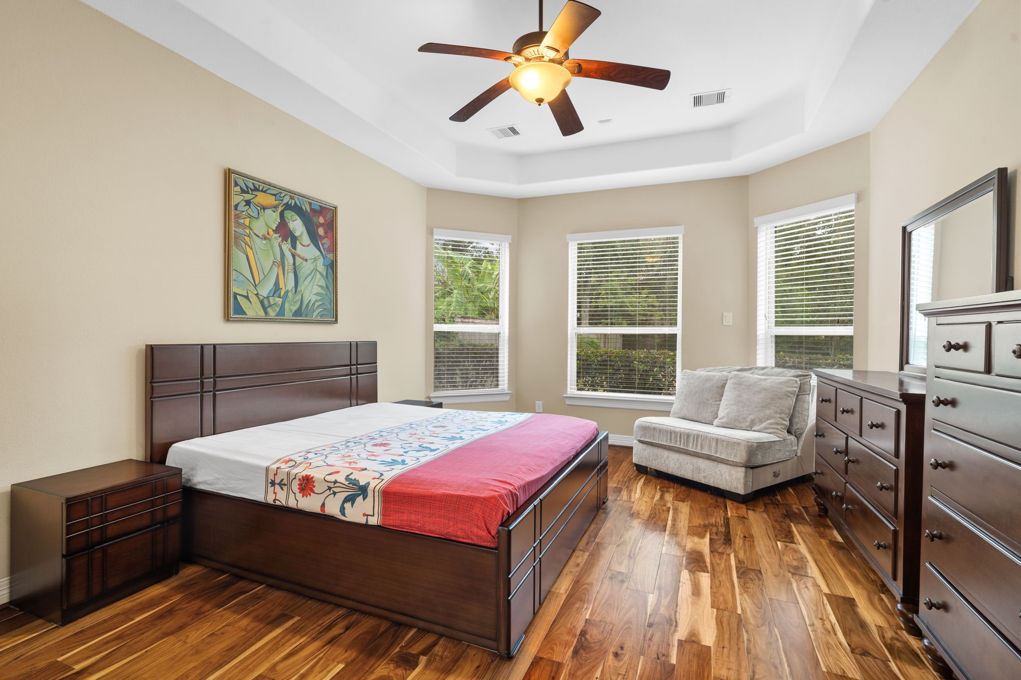 Step inside the spacious, first-floor primary bedroom featuring an elegant tray ceiling, wood flooring, relaxing window views, and an en-suite bathroom.