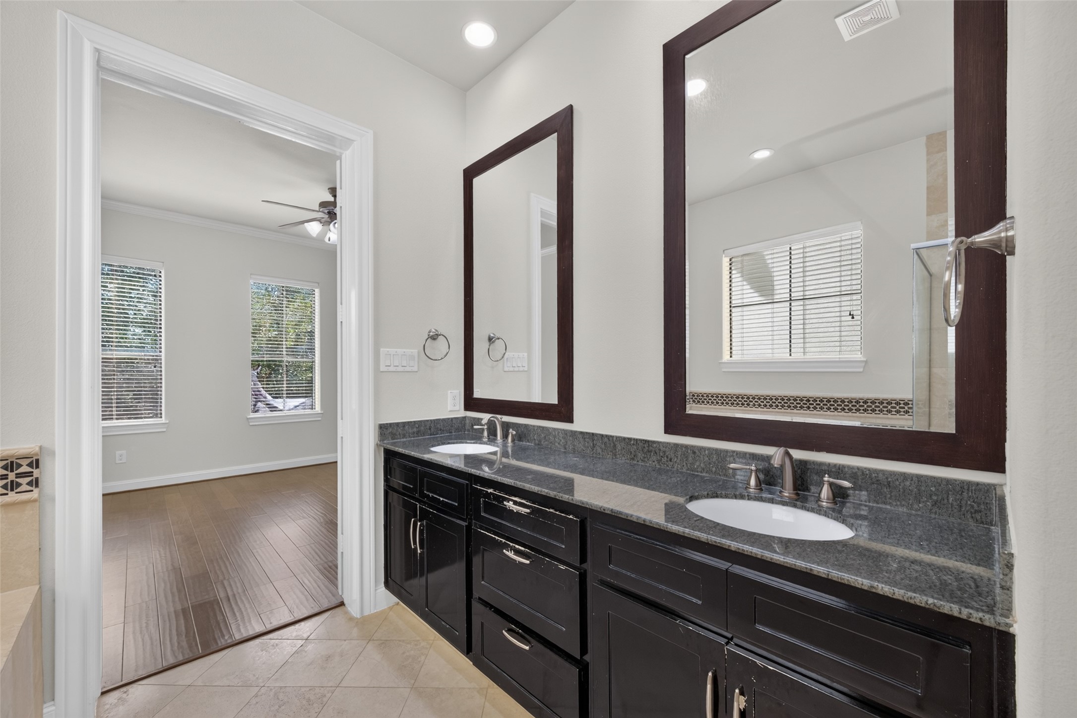 Elegant bathroom featuring a dual-sink vanity with dark cabinetry and granite countertops, opening into a bright room with large windows and hardwood floors.