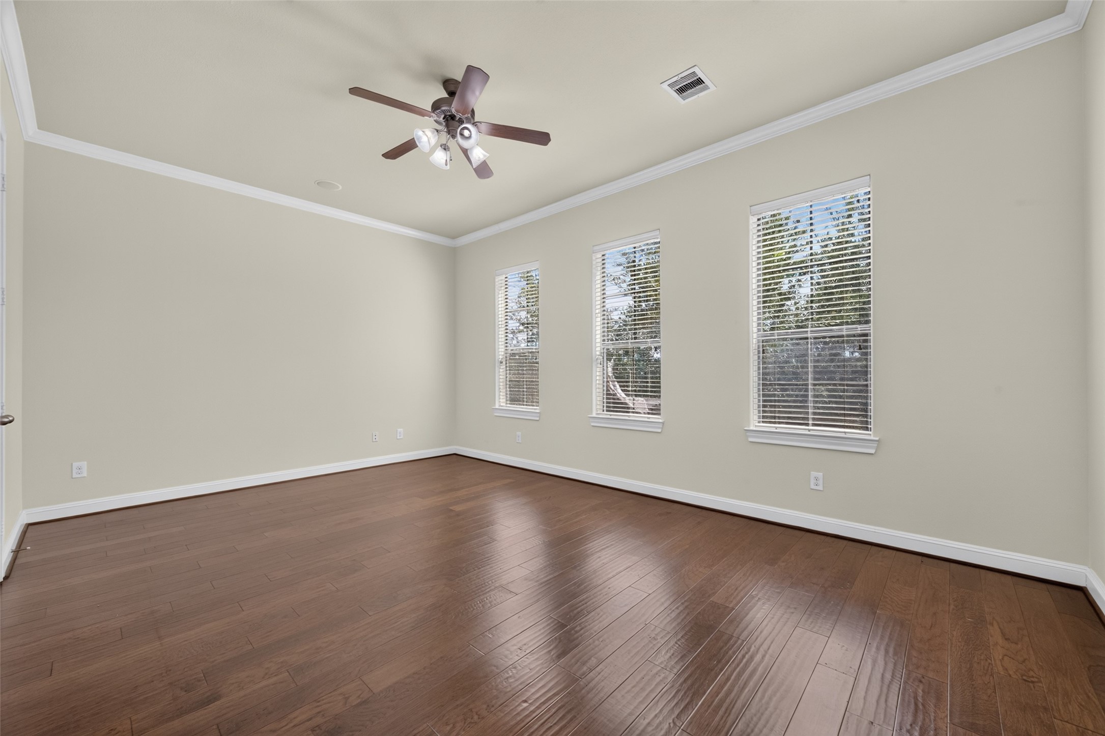 Spacious room with elegant hardwood floors, abundant natural light from three large windows, and a stylish ceiling fan.