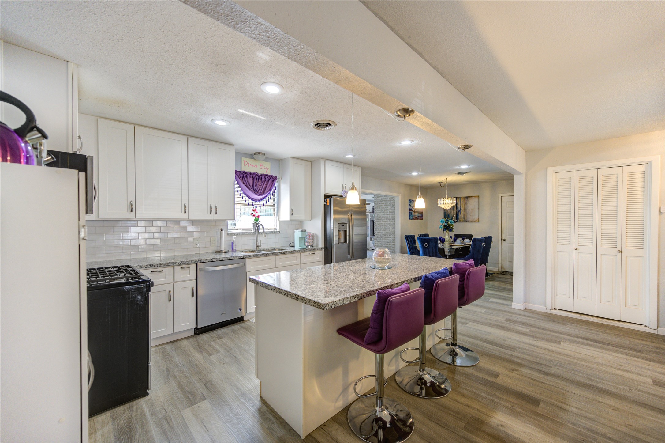 This spacious kitchen features modern white cabinetry, granite countertops, and a large island with bar seating. The open layout connects to a dining area, complemented by elegant lighting and wood-style flooring. Perfect for entertaining and family gatherings.