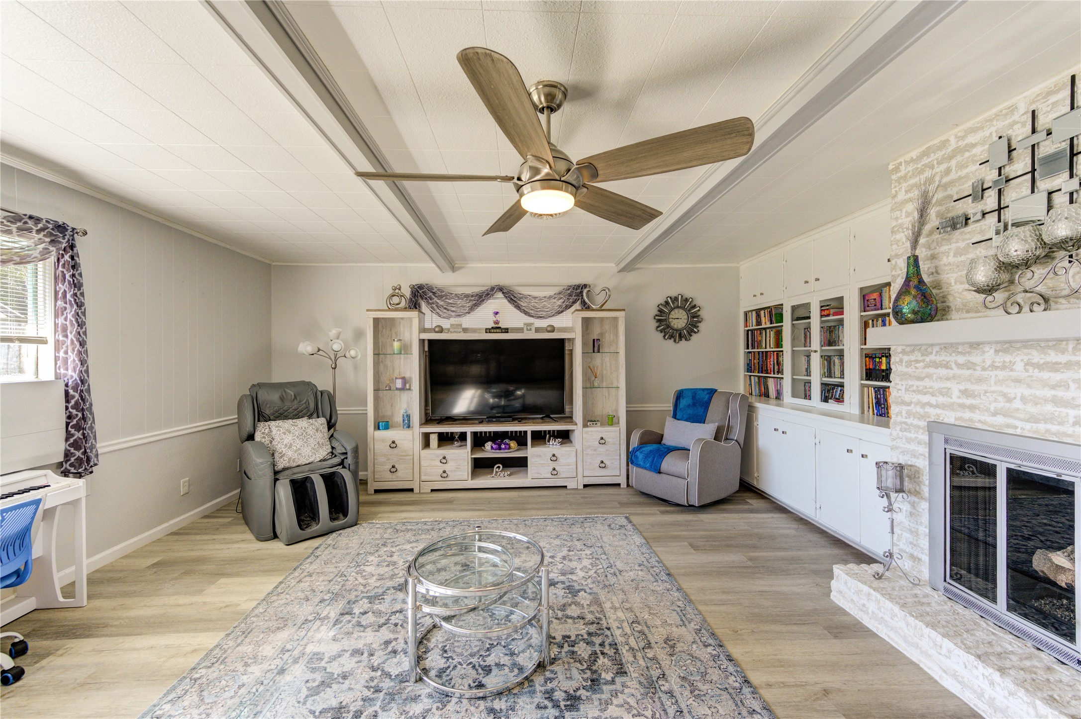 The den features a modern ceiling fan, built-in bookshelves, and a white painted brick fireplace. Great natural light shines through the large windows.