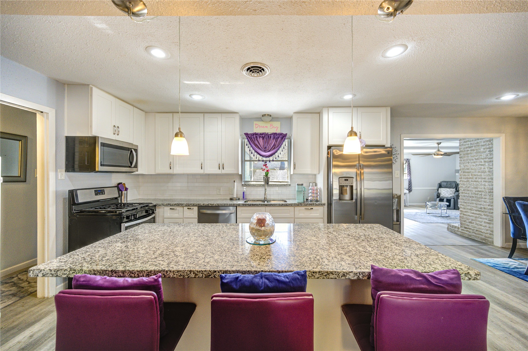 This modern kitchen features granite countertops, a large island with bar seating, stainless steel appliances, and white cabinetry. It opens into a spacious living area, creating a seamless flow for entertaining.