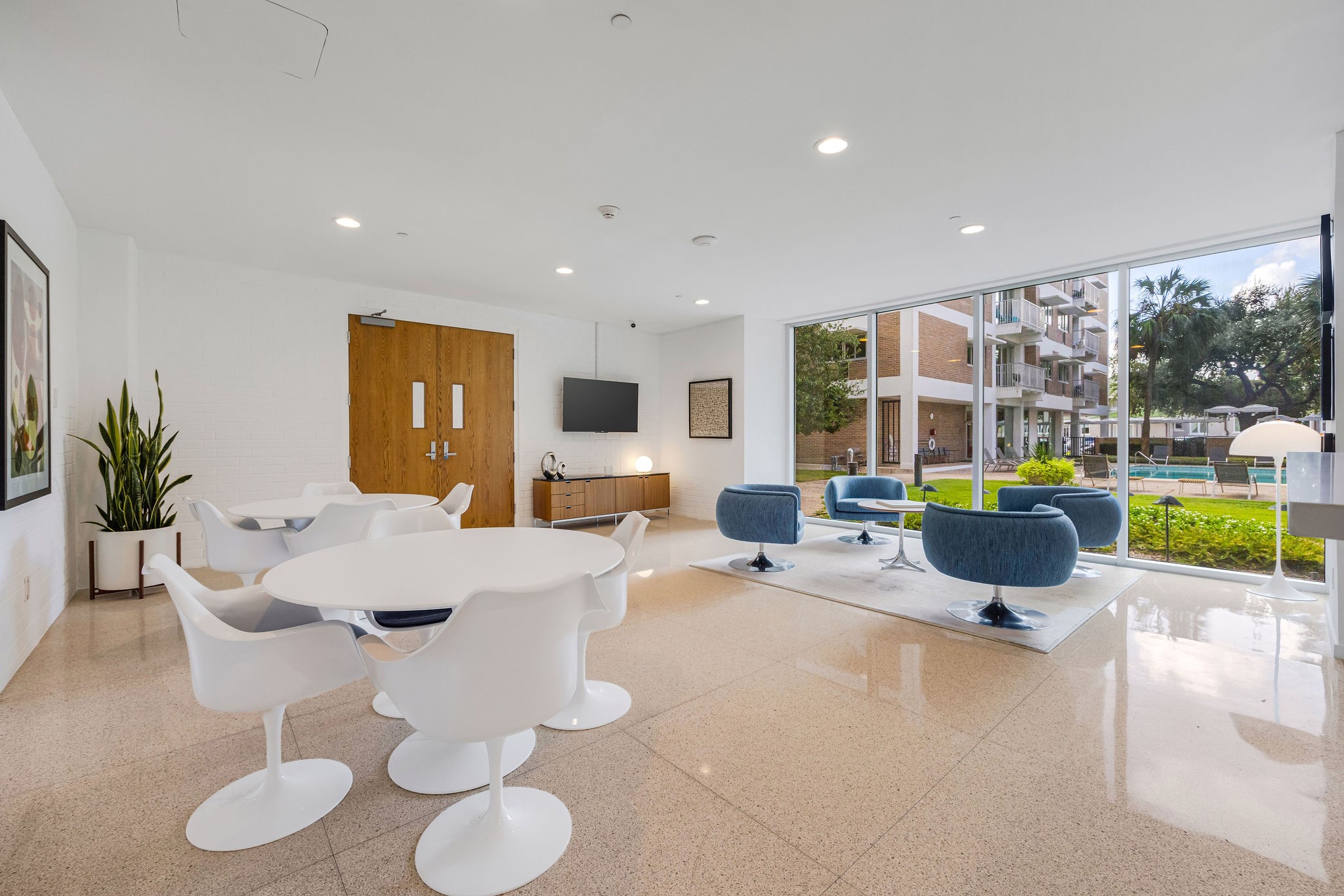 Bright and modern communal space featuring sleek white and blue seating, polished floors, and large windows offering views of a lush courtyard and pool area.