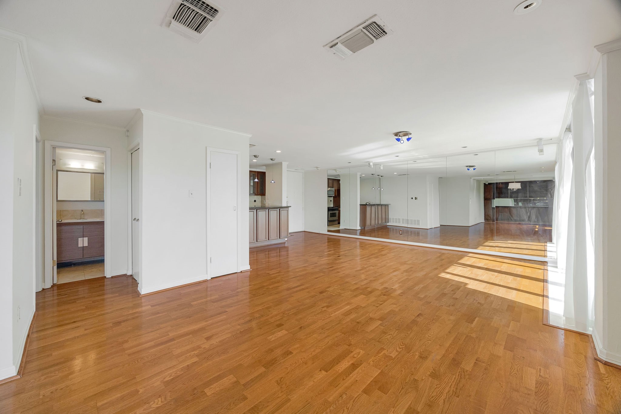 Spacious living area with gleaming hardwood floors and abundant natural light, featuring a mirrored wall and open layout that flows into a modern kitchen space.
