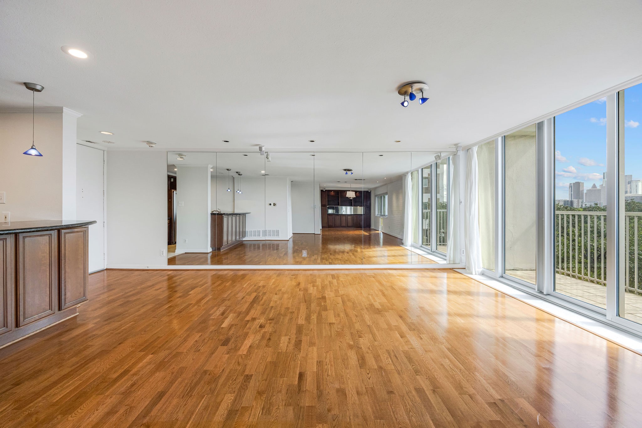 Spacious open-concept living area with gleaming hardwood floors, floor-to-ceiling windows offering abundant natural light and stunning city views, complemented by a mirrored accent wall and modern kitchen cabinetry.