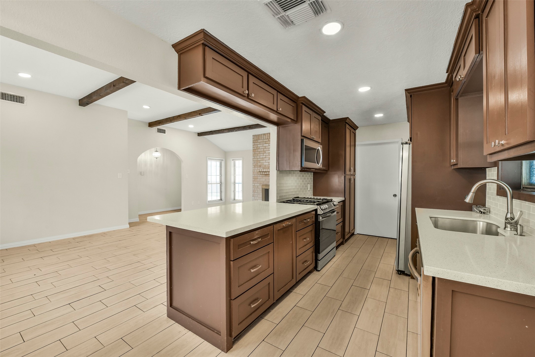 Rich wooden cabinetry, modern stainless steel appliances, and sleek countertops, seamlessly opening to a bright living area with stylish wood-look tile flooring and exposed beams.