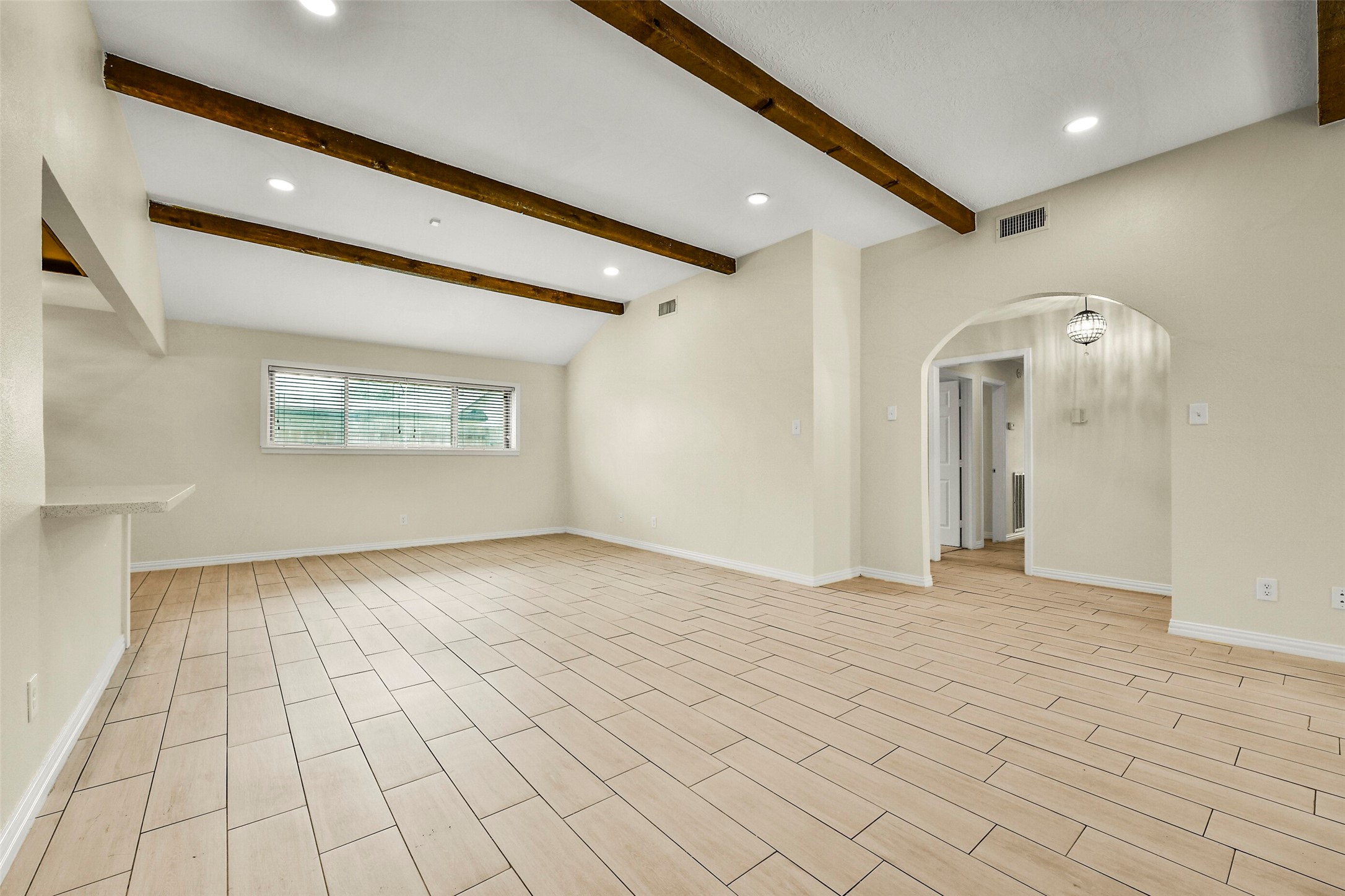 Spacious living area with elegant wooden beams, sleek tile flooring, and abundant natural light from a large window, leading into an adjoining room through an arched doorway. Spacious living area with elegant wooden beams, sleek tile flooring, and abundant natural light from a large window, leading into an adjoining room through an arched doorway.