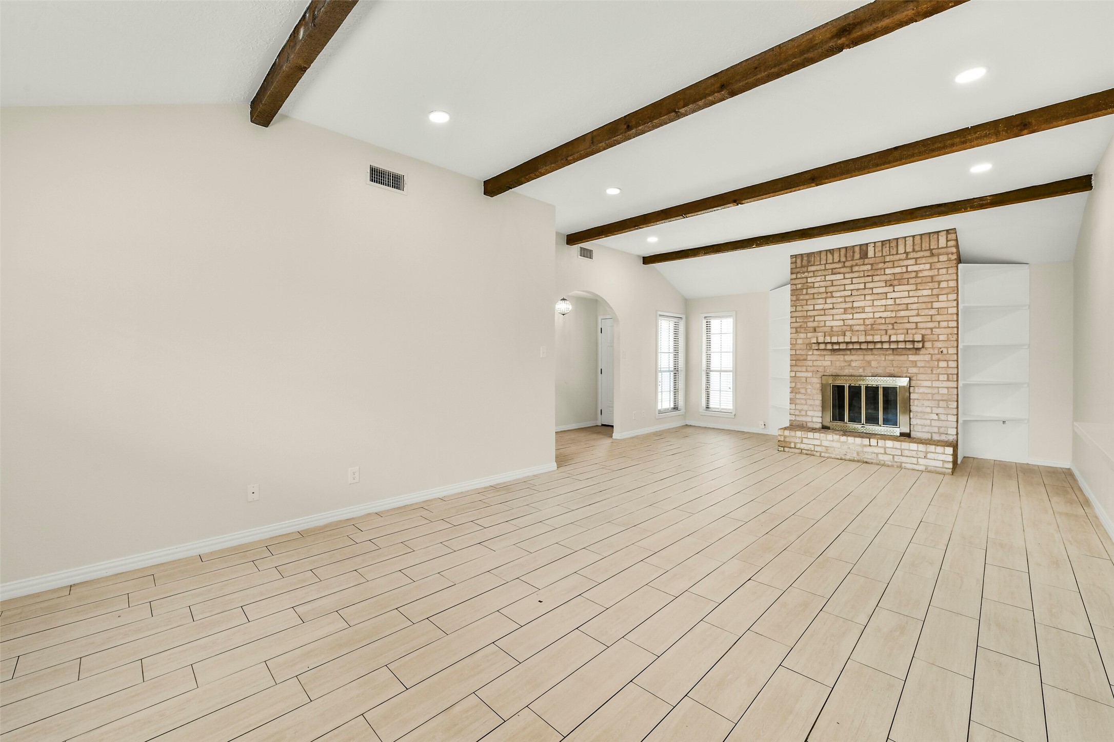 Living area featuring elegant wood-look tile flooring, exposed wood beams, and a charming brick fireplace, perfect for cozy gatherings. Living area featuring elegant wood-look tile flooring, exposed wood beams, and a charming brick fireplace, perfect for cozy gatherings.