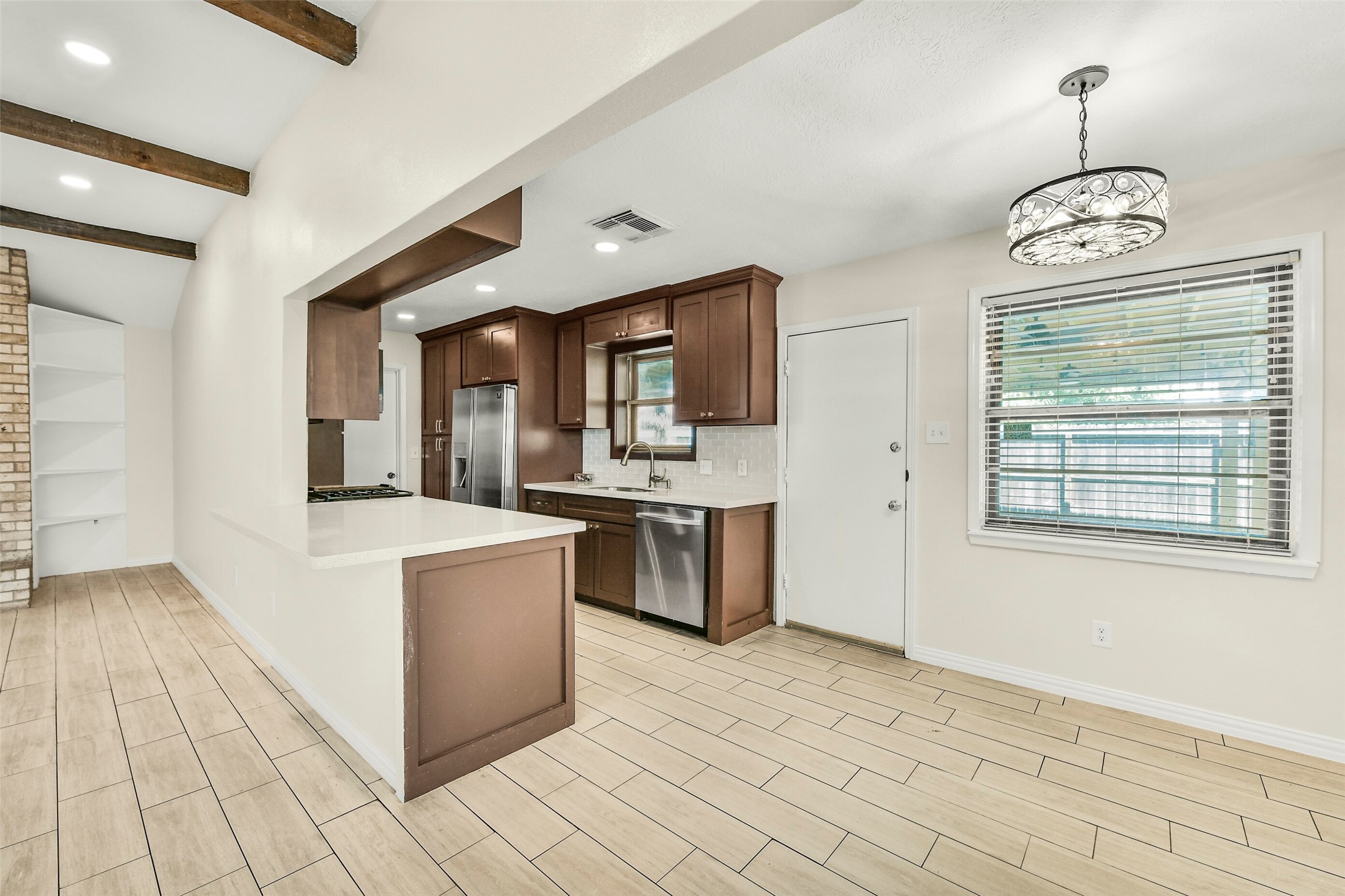 Kitchen with sleek stainless steel appliances, rich wood cabinetry, and a spacious breakfast bar. The open layout flows into the living area, highlighted by exposed beams and a cozy brick accent wall with built-in shelving. Natural light pours in through the large window, enhancing the inviting atmosphere.