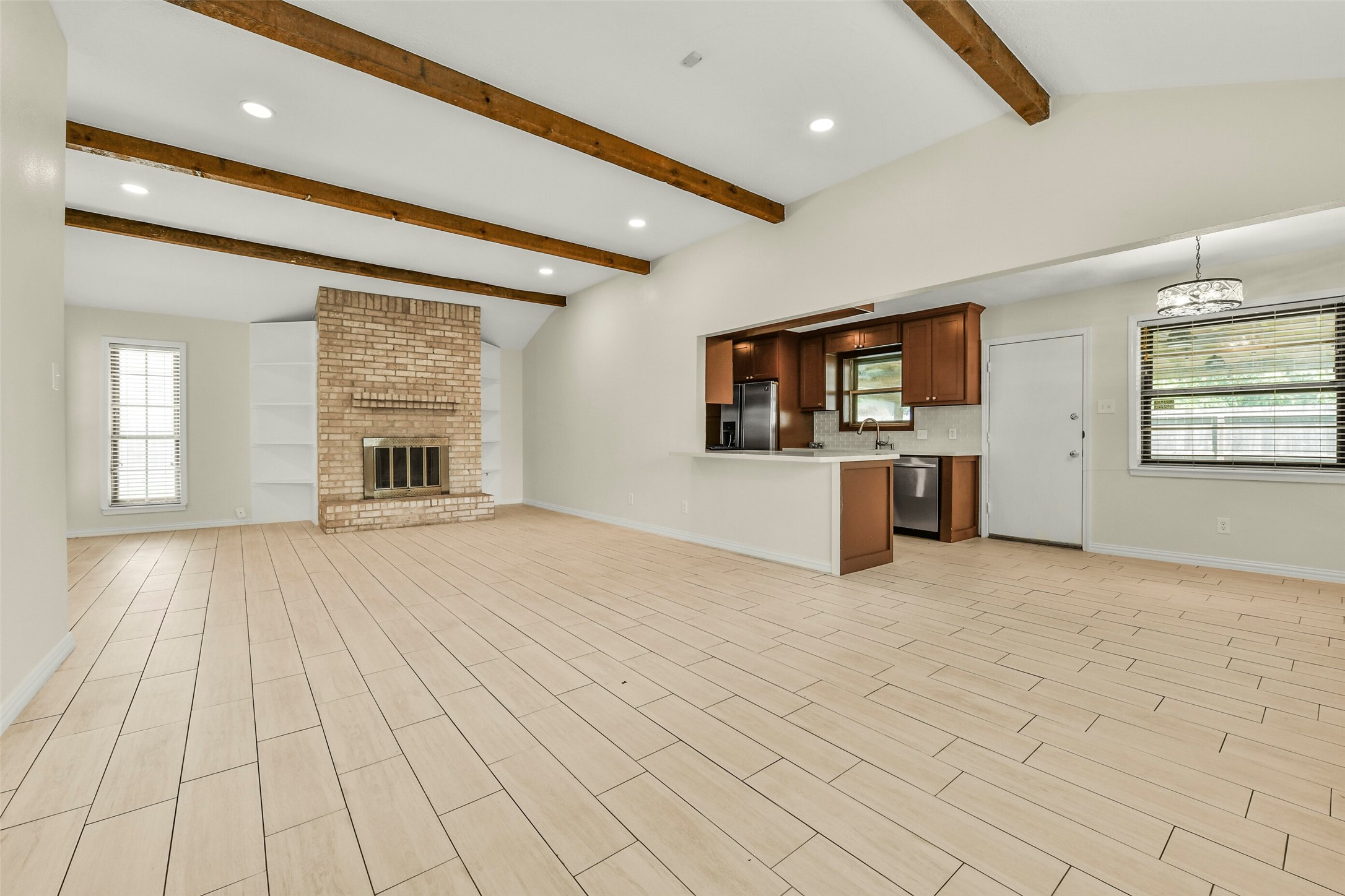 Spacious open-concept living area features elegant wood-beam ceilings, a cozy brick fireplace, and sleek tile flooring, seamlessly connecting to a modern kitchen with stainless steel appliances and warm cabinetry. Spacious open-concept living area features elegant wood-beam ceilings, a cozy brick fireplace, and sleek tile flooring, seamlessly connecting to a modern kitchen with stainless steel appliances and warm cabinetry.