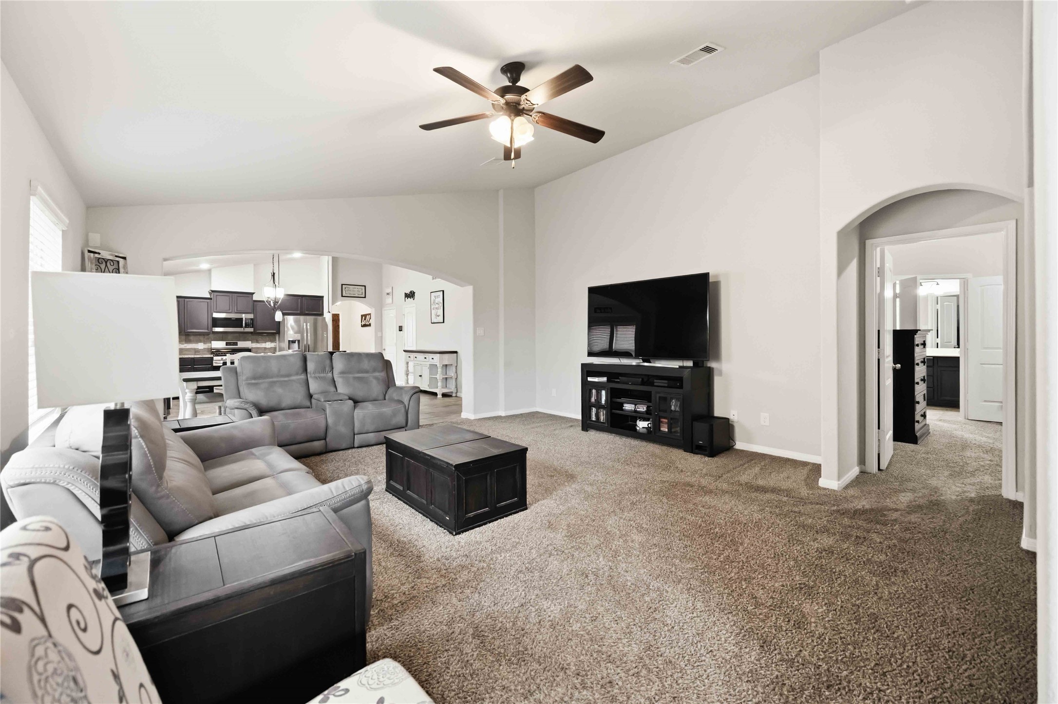 Spacious living room with plush carpeting, a ceiling fan, and modern furnishings, seamlessly connected to an open-concept kitchen and dining area. Spacious living room with plush carpeting, a ceiling fan, and modern furnishings, seamlessly connected to an open-concept kitchen and dining area.