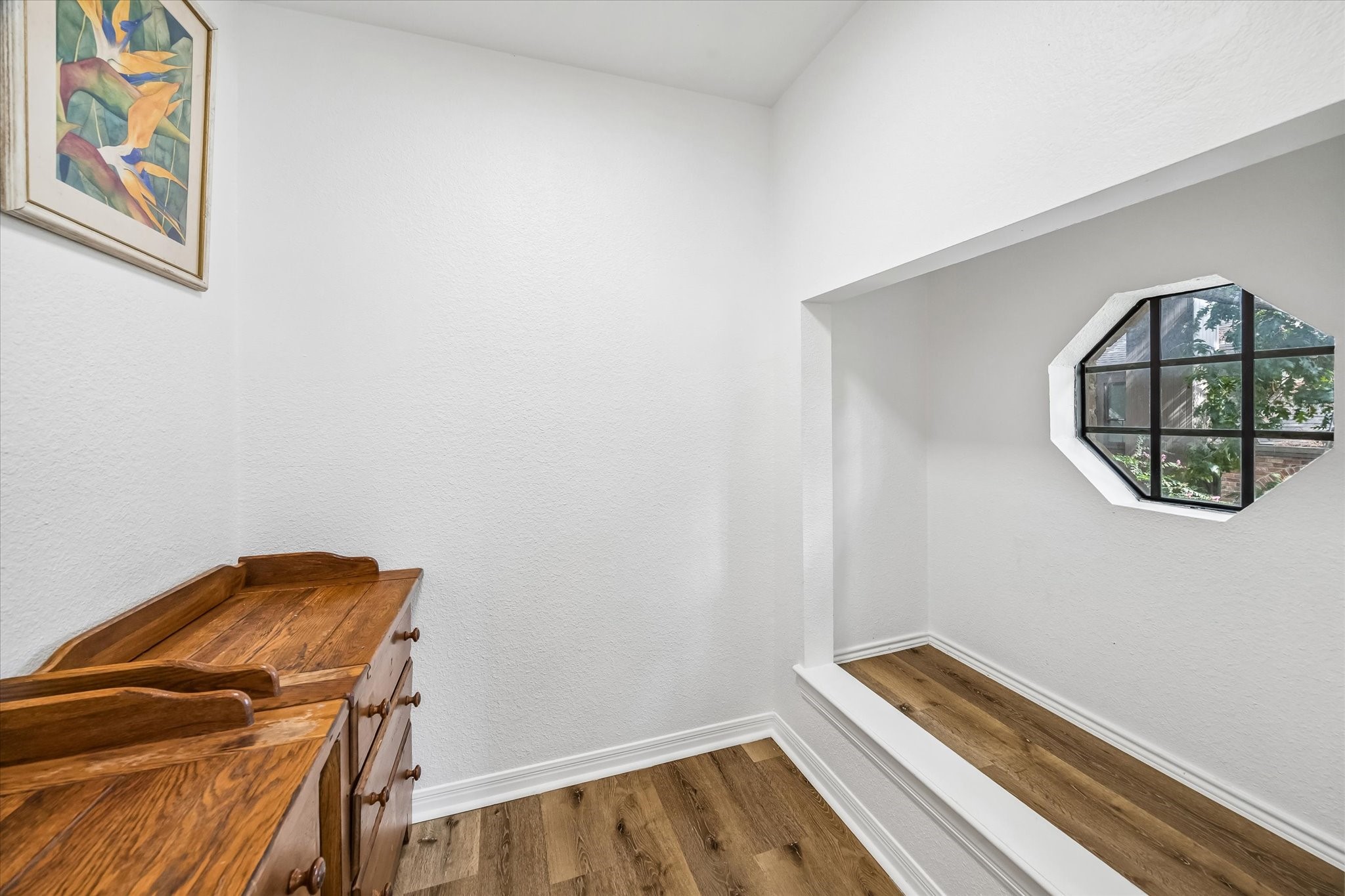 Charming nook with a unique octagonal window, offering natural light and complemented by warm hardwood flooring and a cozy wooden cabinet.