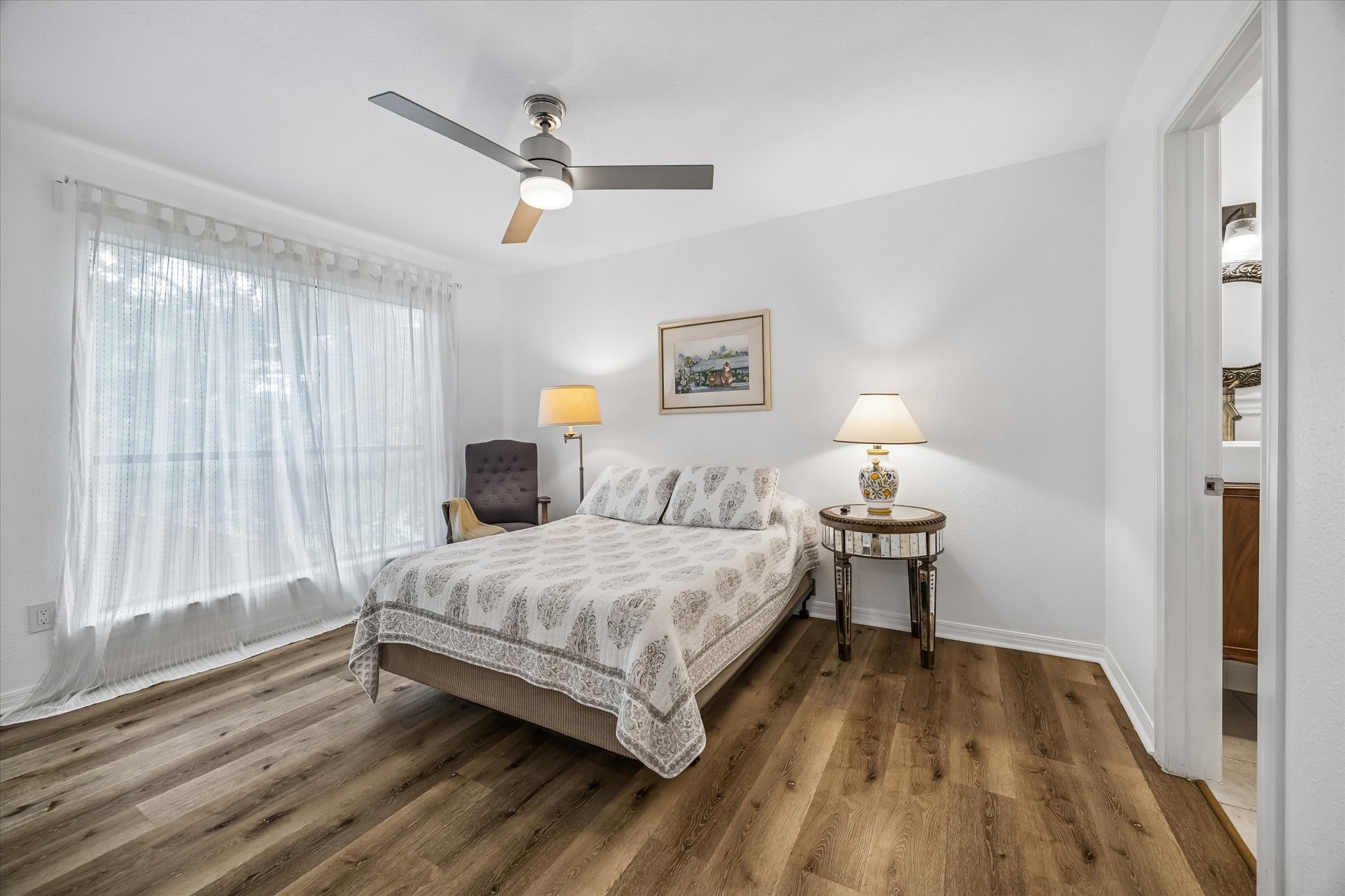 Bright and inviting bedroom featuring a large window with sheer curtains, new vinyl plank flooring, and a ceiling fan for comfort.