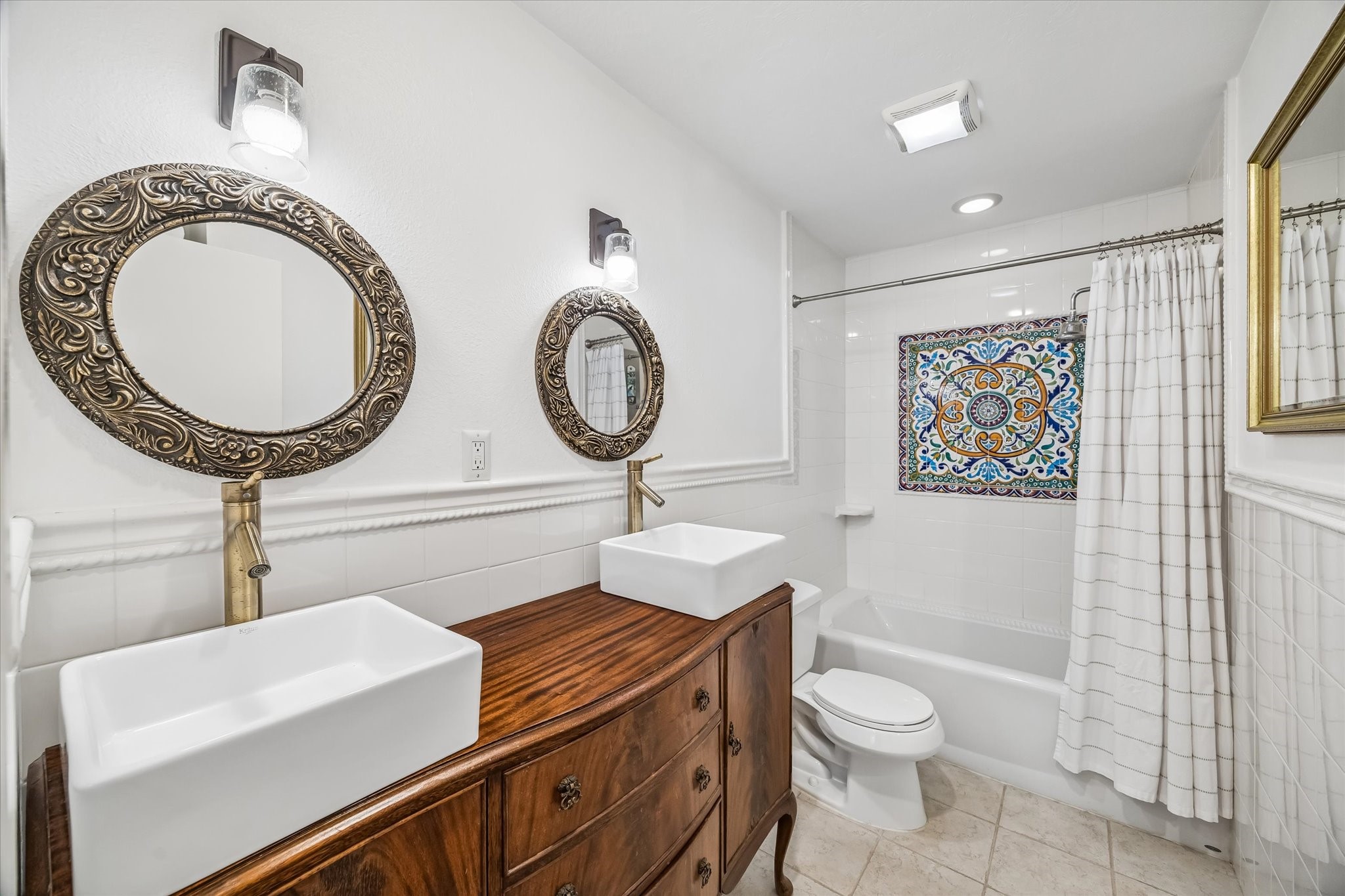 Elegant bathroom featuring dual vessel sinks on a freshly restored vintage wooden vanity, ornate mirrors, and a beautifully tiled shower with a colorful mosaic accent.