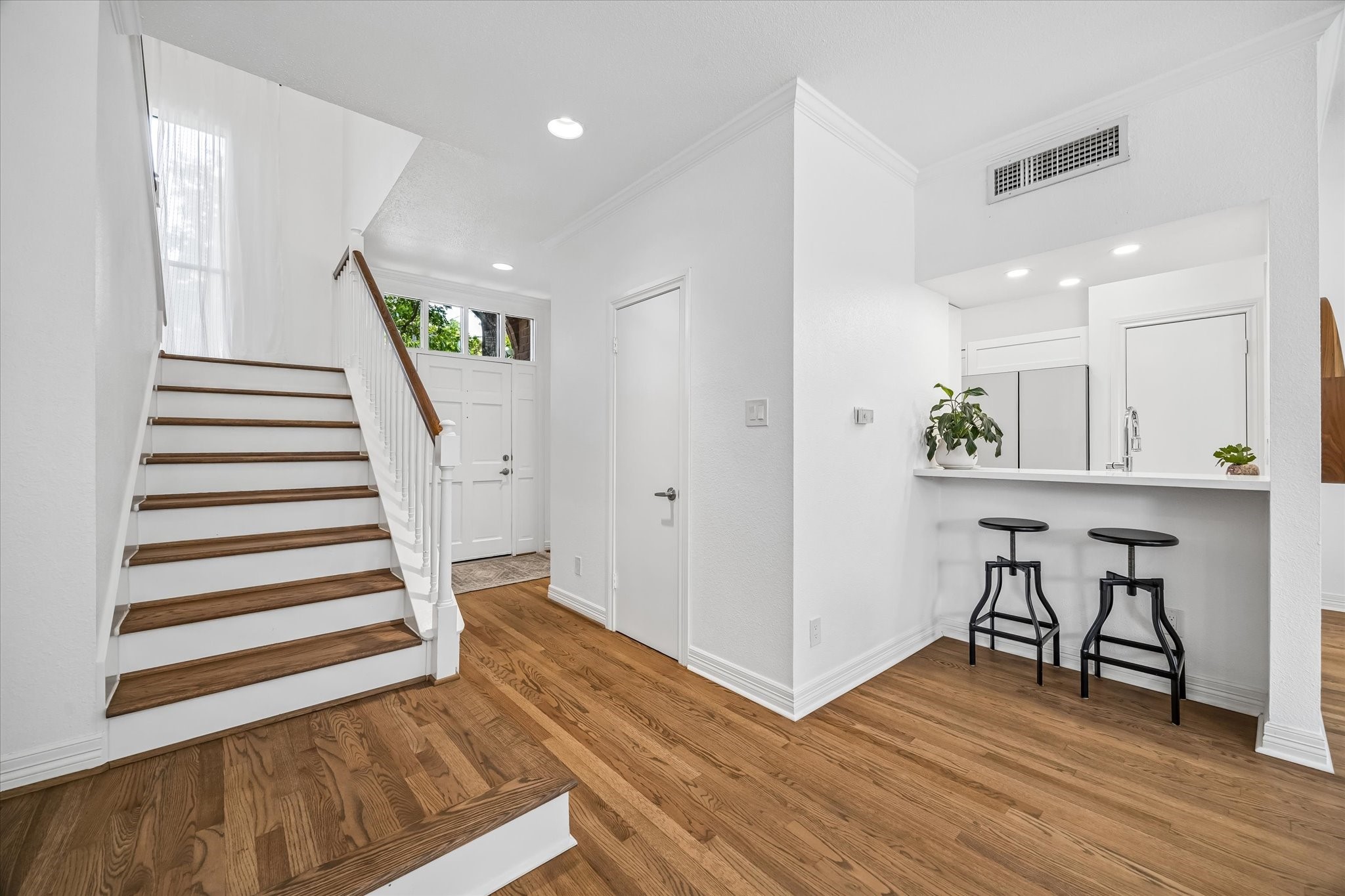 Bright and inviting entryway featuring elegant hardwood floors, a modern staircase, and a cozy breakfast bar leading to a well-lit kitchen area.