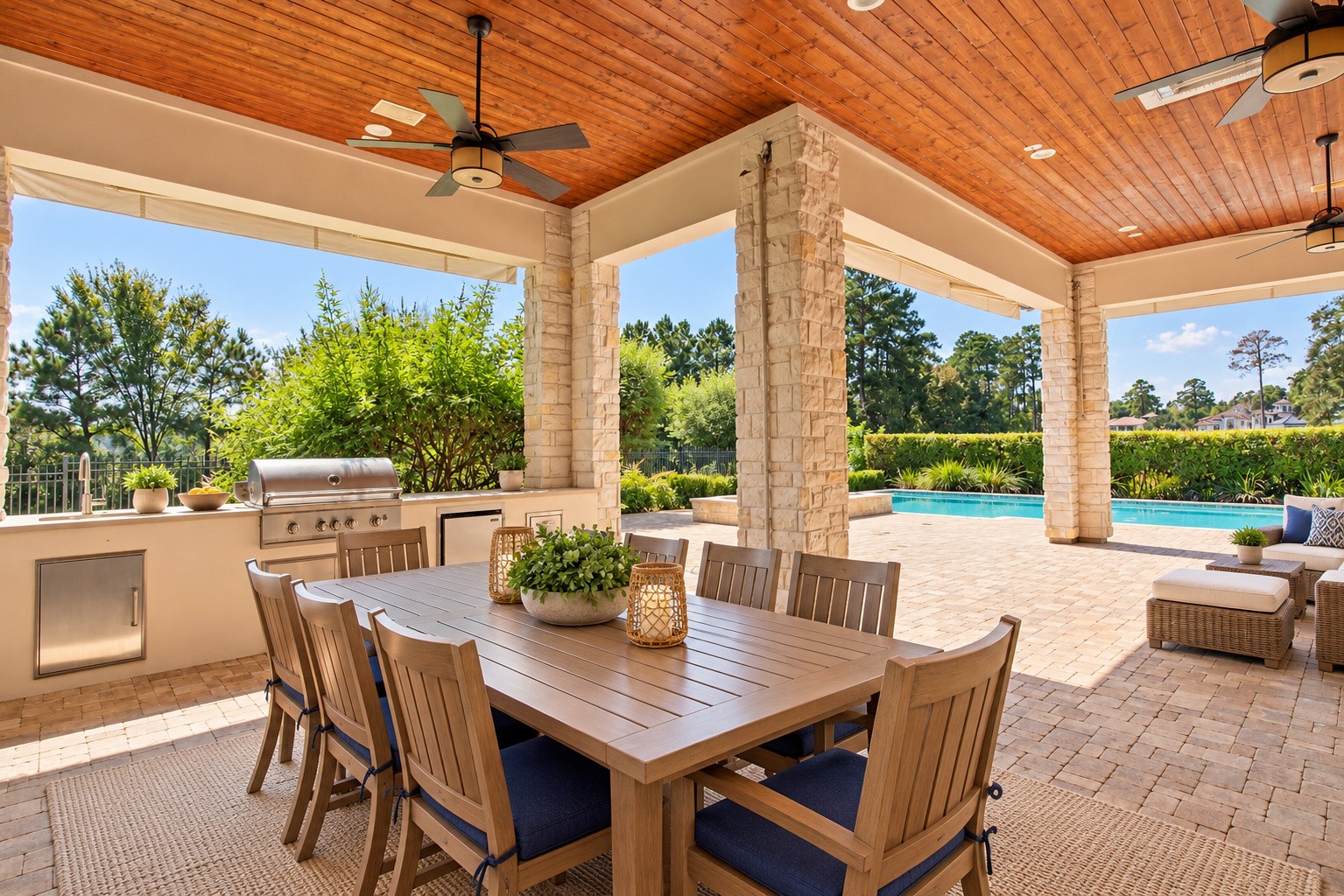 Impressive outdoor dining area featuring a rich wood-paneled ceiling, beautiful stone columns, and a fully equipped summer kitchen overlooking the pool.