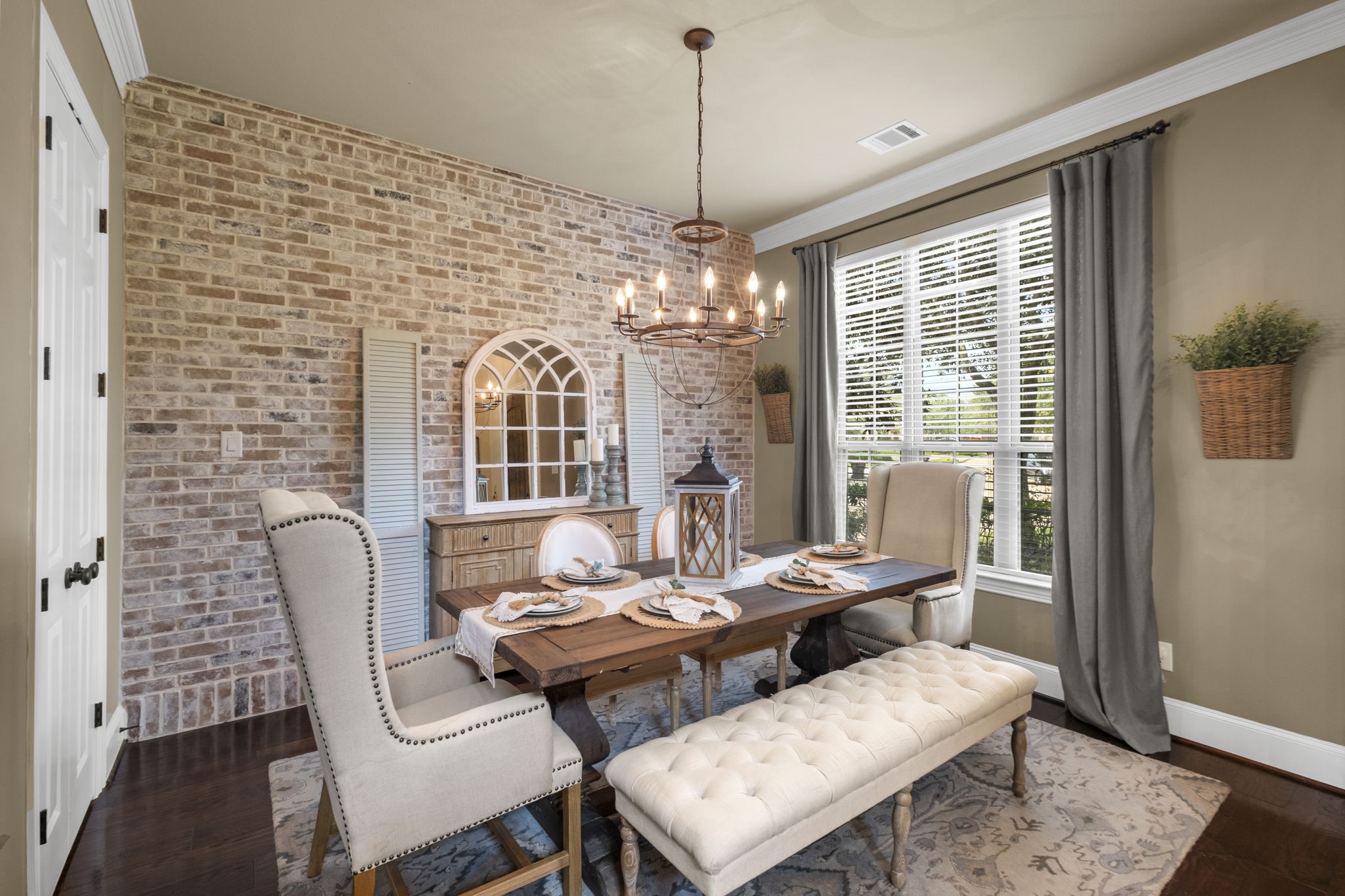 Formal dining area highlighting an accent wall made of exposed brick finish, detailed crown molding, rich hardwood floors, and a wrought-iron candle-style chandelier. Large windows with custom blinds and drapery fill the space with natural light.