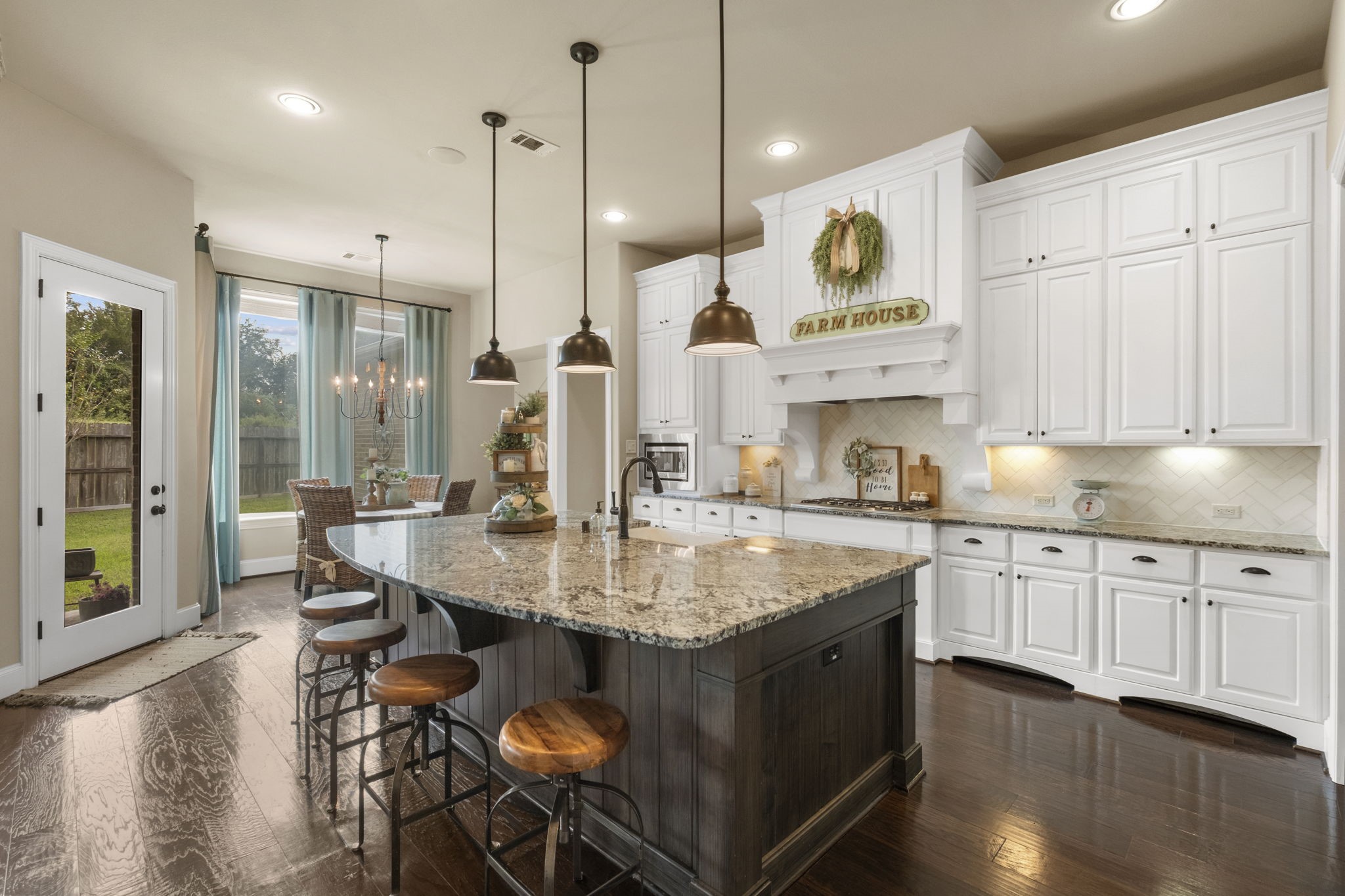 Gourmet kitchen showcasing custom white cabinetry, dark hardwood floors, and a massive dark-stained contrasting center island with granite countertops and pendant lighting. Includes a decorative white range hood with tile herringbone backsplash, undermount lighting, and seamless flow into the breakfast nook.