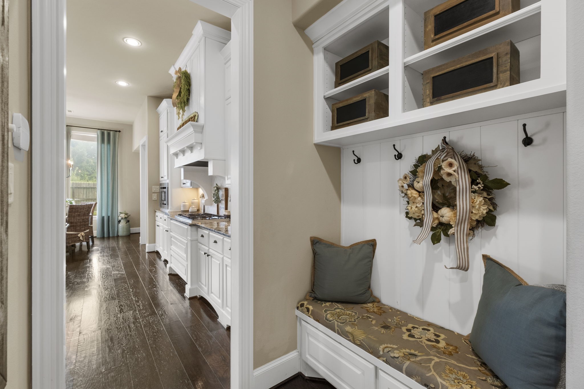 Custom built-in mudroom entry nook featuring beadboard accent paneling, storage coat hooks, upper display cubbies, and a fitted bench with built-in lower drawers. Hallway beyond leads directly into the main kitchen area.