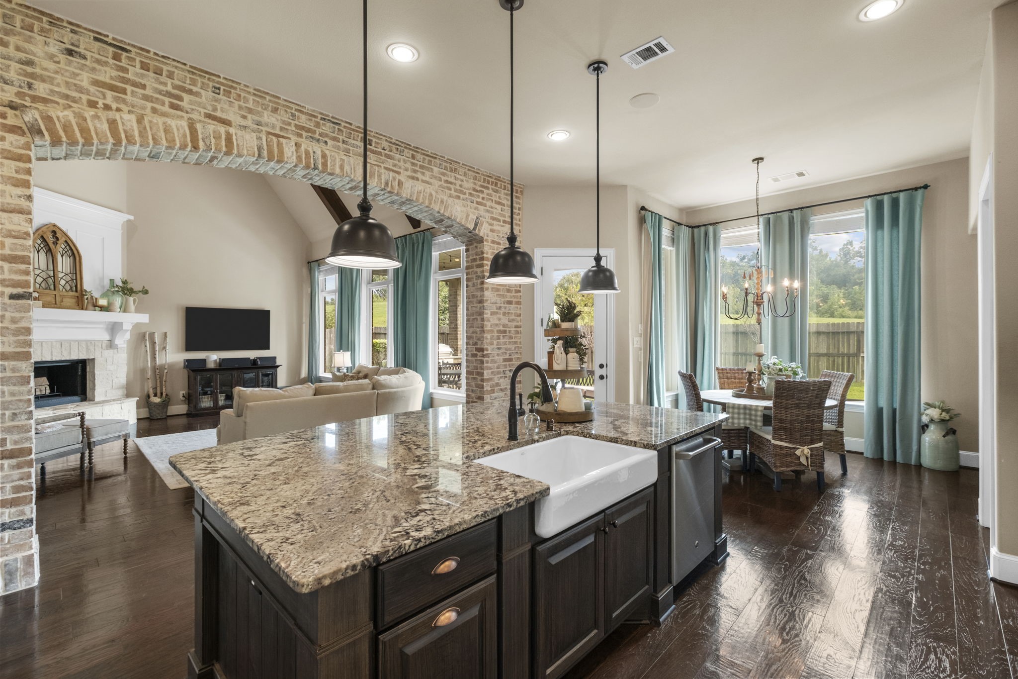 View from the kitchen island featuring an apron-front farmhouse sink with a gooseneck faucet and built-in dishwasher. Showcases the dramatic exposed brick archway separating the kitchen from the living area, along with the adjacent casual breakfast nook.