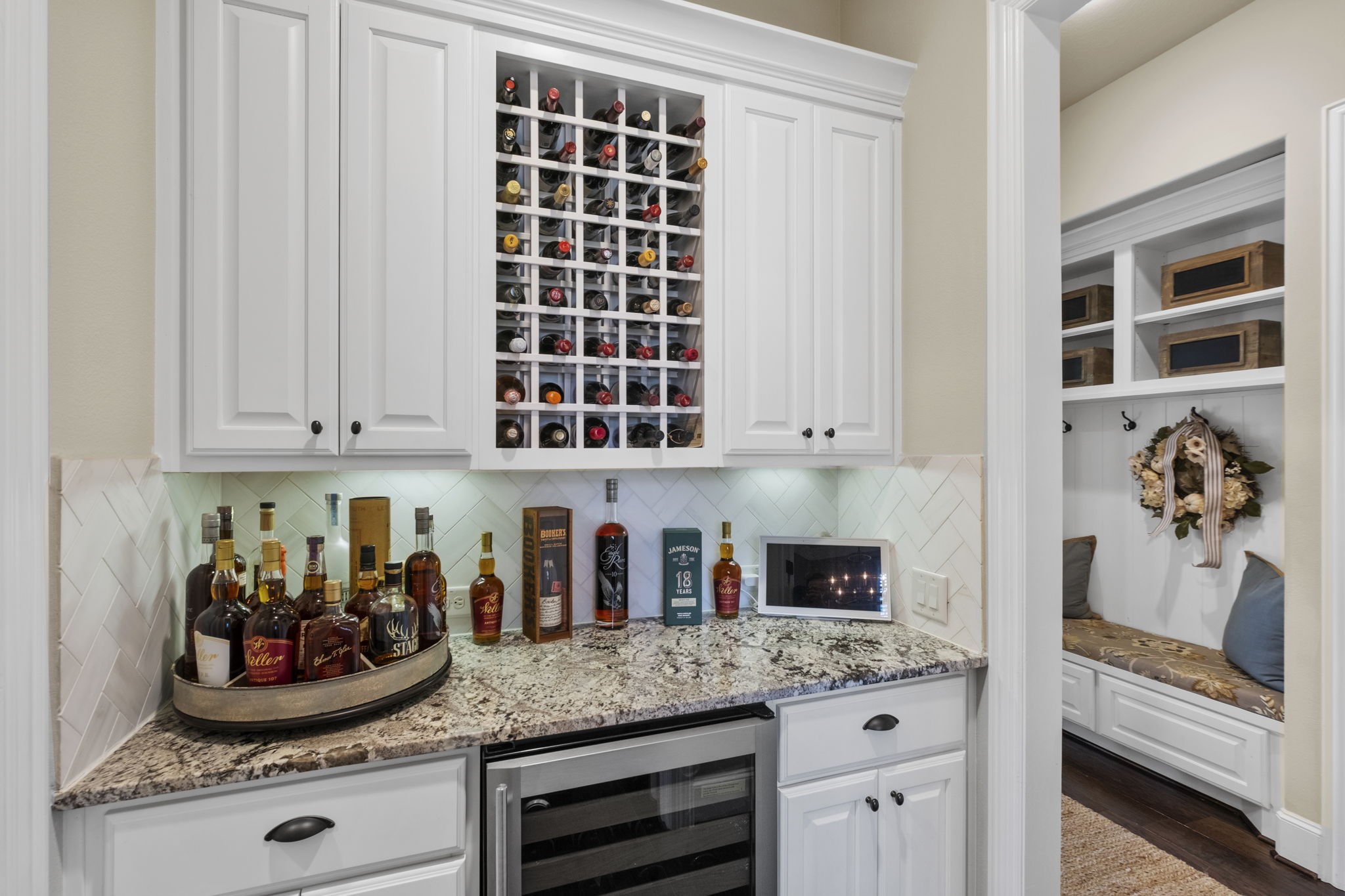 Dedicated built-in dry bar area boasting a custom white cabinet surround, integrated built-in wine rack lattice, granite countertop, herringbone tile backsplash, under-cabinet lighting, and space for a beverage cooler.